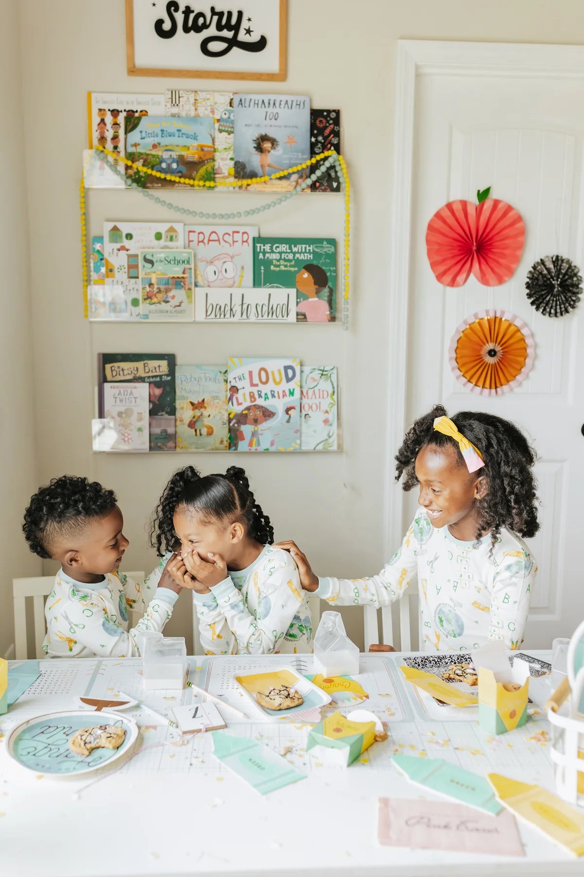 Three children in pajamas laughing at a back-to-school breakfast table, surrounded by books, themed party decor, and paper fans on the wall and door.