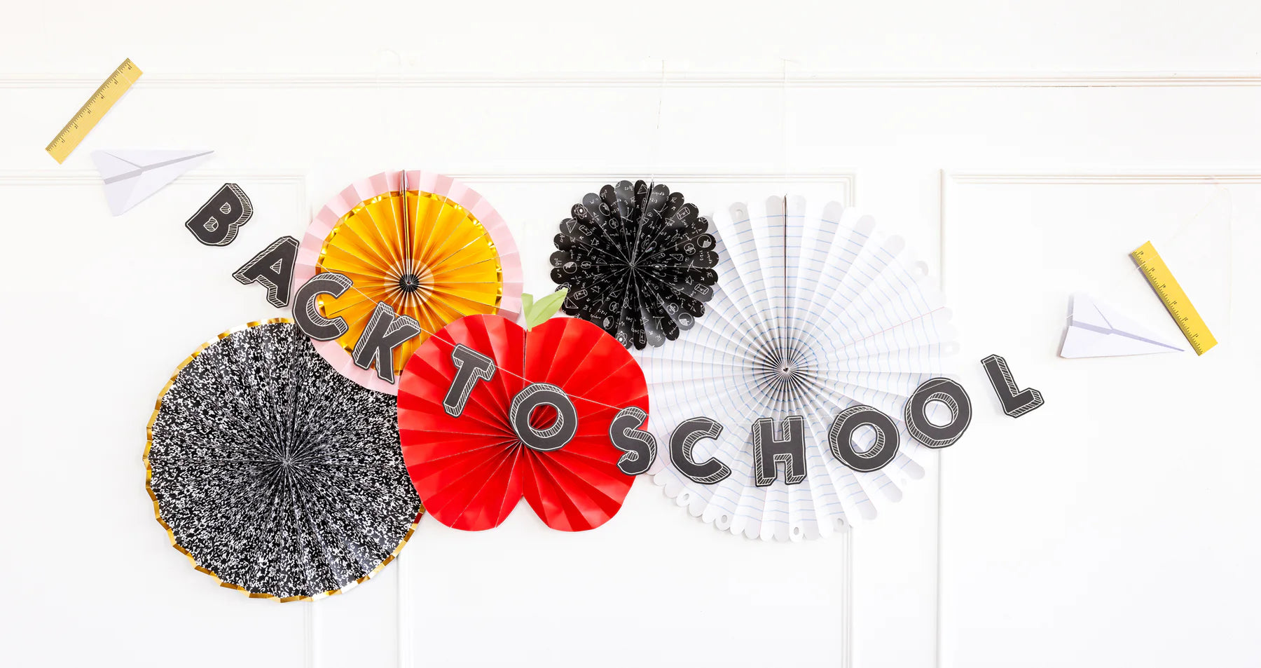 Back-to-school party setup featuring a “Back to School” banner over a wall decorated with themed paper fans and school ruler and paper airplane cutouts.