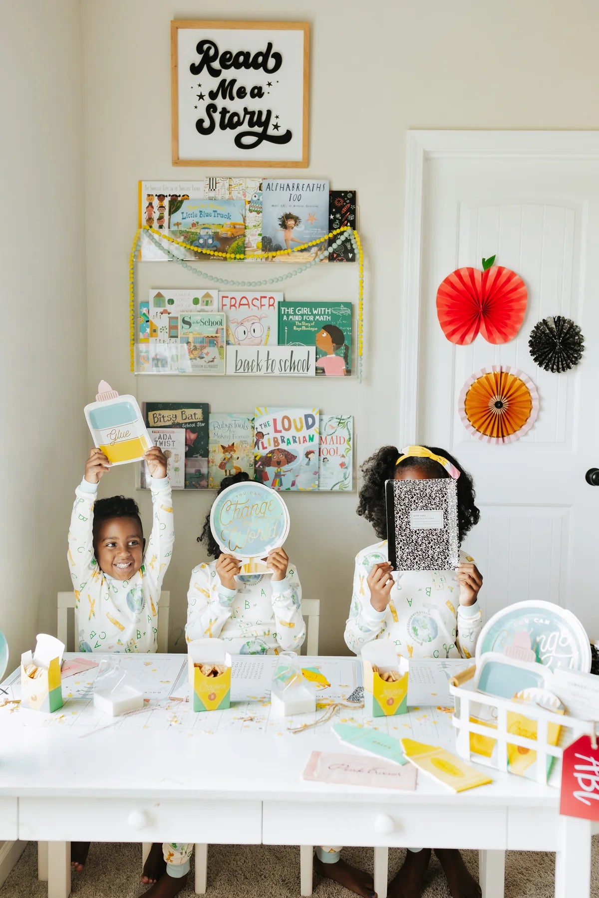 Three young children sitting at a decorated table holding up school-themed party plates and notebooks.
