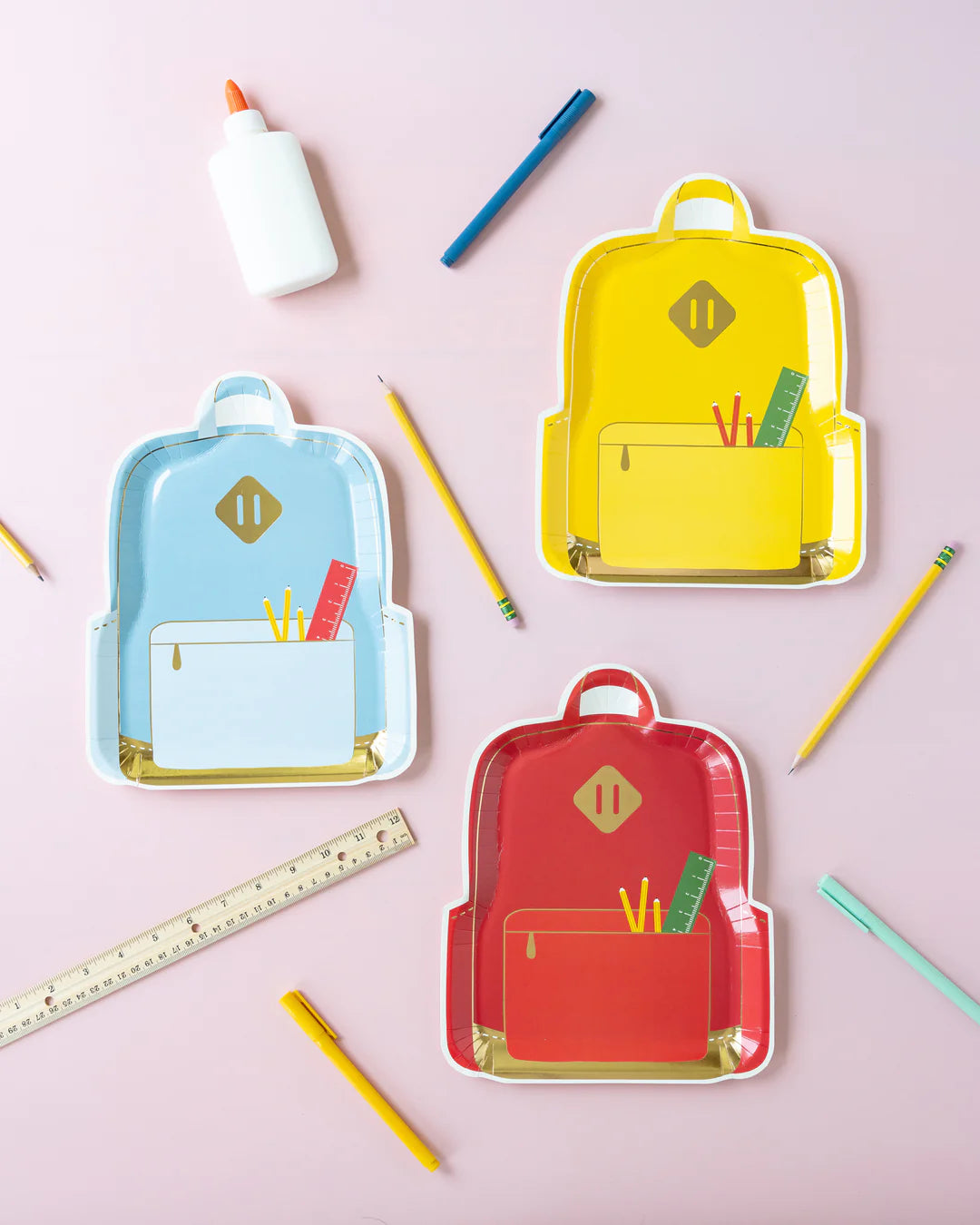 Flat lay of three backpack plates in red, yellow, and blue surrounded by school supplies like pencils, glue, and rulers