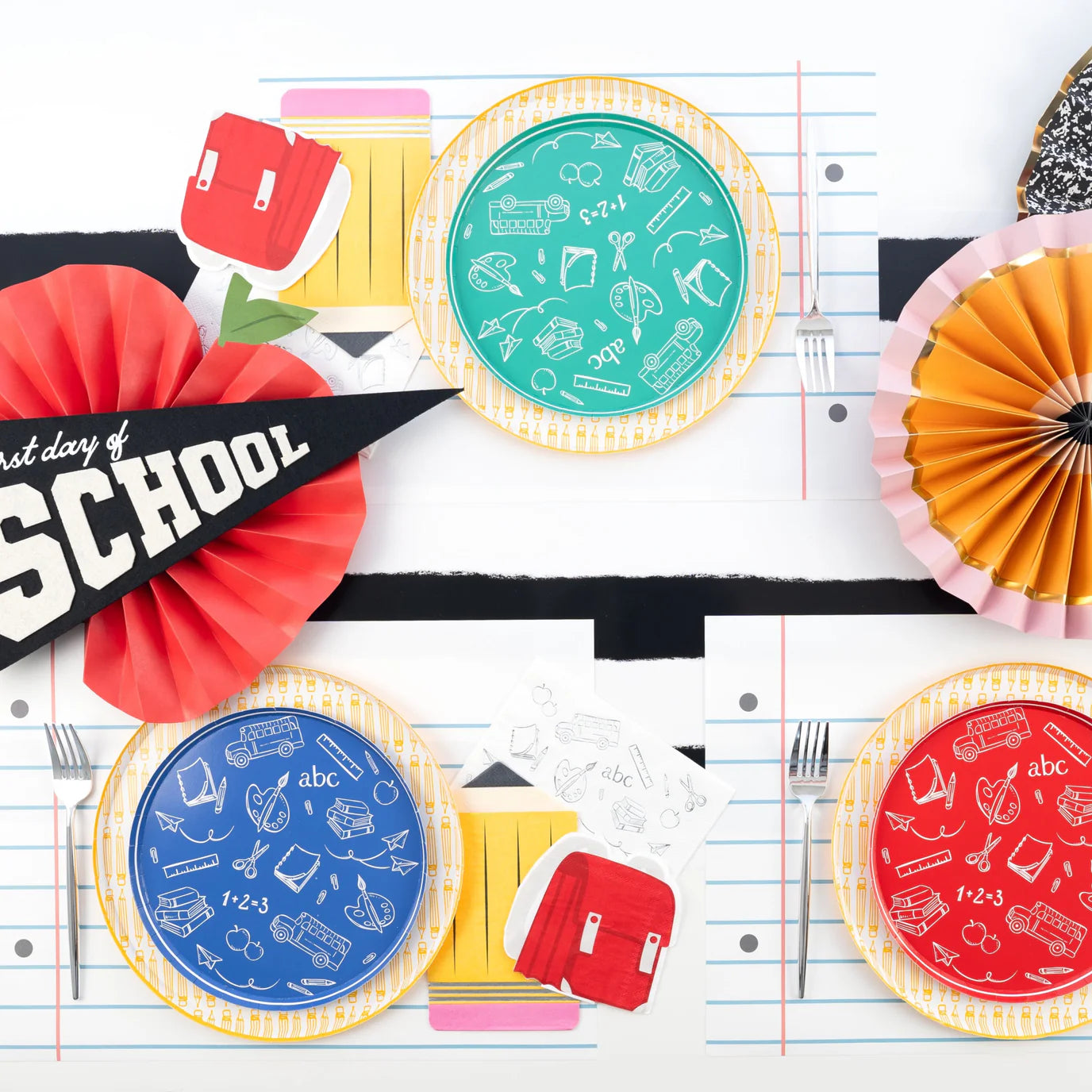 Notebook‑placemat tablescape: green, blue, red icon plates with pencil napkin, red ‘School’ pennant.