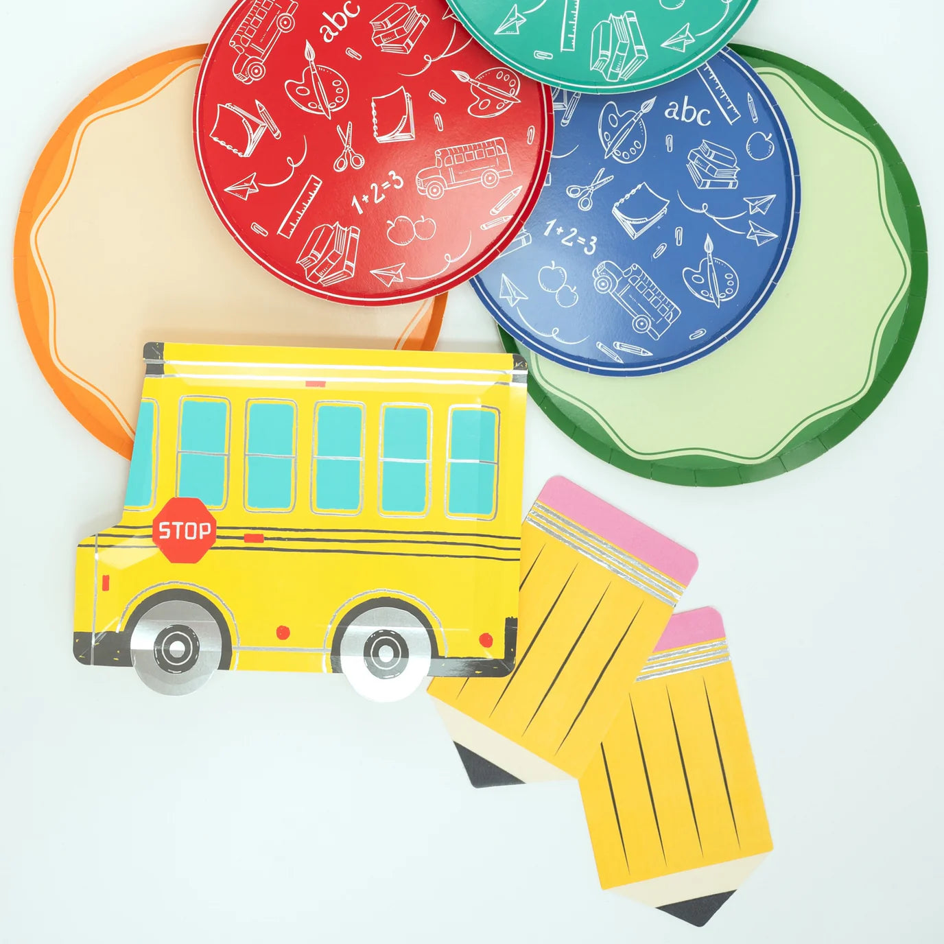 Overhead view of colorful school‑themed plates, yellow school‑bus plate, and pencil napkins fanned out on white background