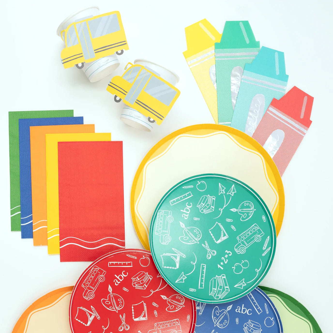 Overhead flat lay of colorful school‑themed tableware: yellow bus cups, rainbow napkins, pencil dinner plates, and green/red/blue icon plates on white background.