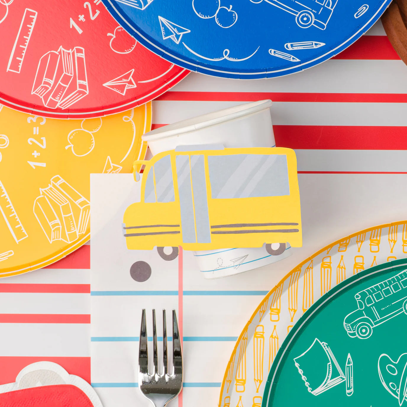 Close‑up of bus cup next to yellow pencil dinner plate, fork, and red/yellow school‑icon plates on striped runner.
