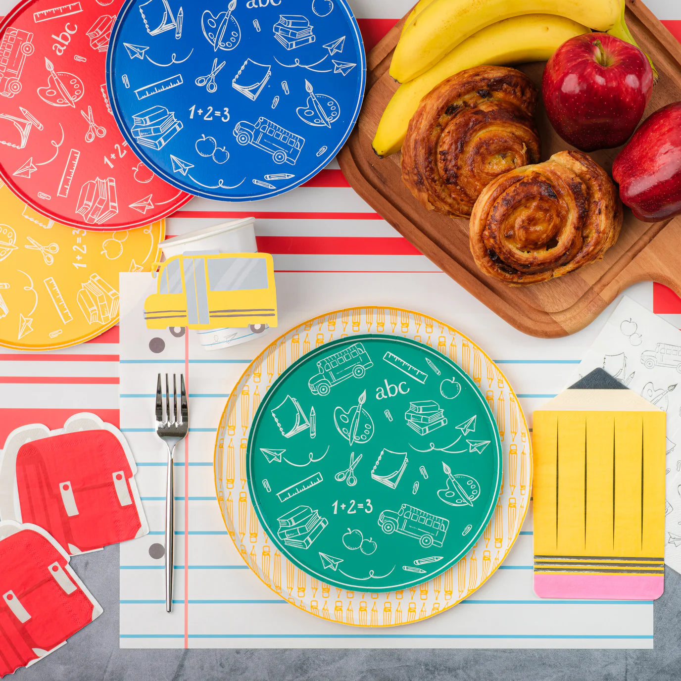 Breakfast scene: pastries, bananas, green icon plate on pencil plate, bus cup, pencil napkin, and notebook placemat over red stripes.