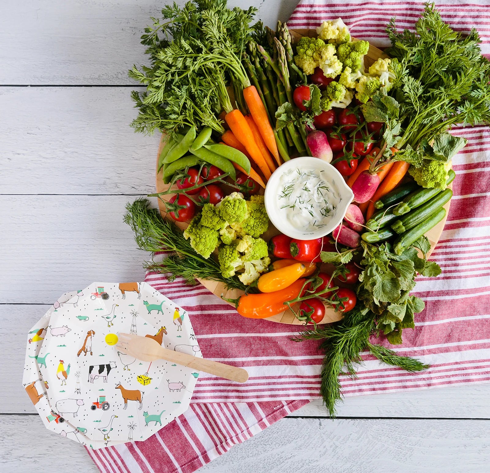 Wood table set with farm animal dessert plates, wooden cutlery, veggie platter, and striped red tea towel.