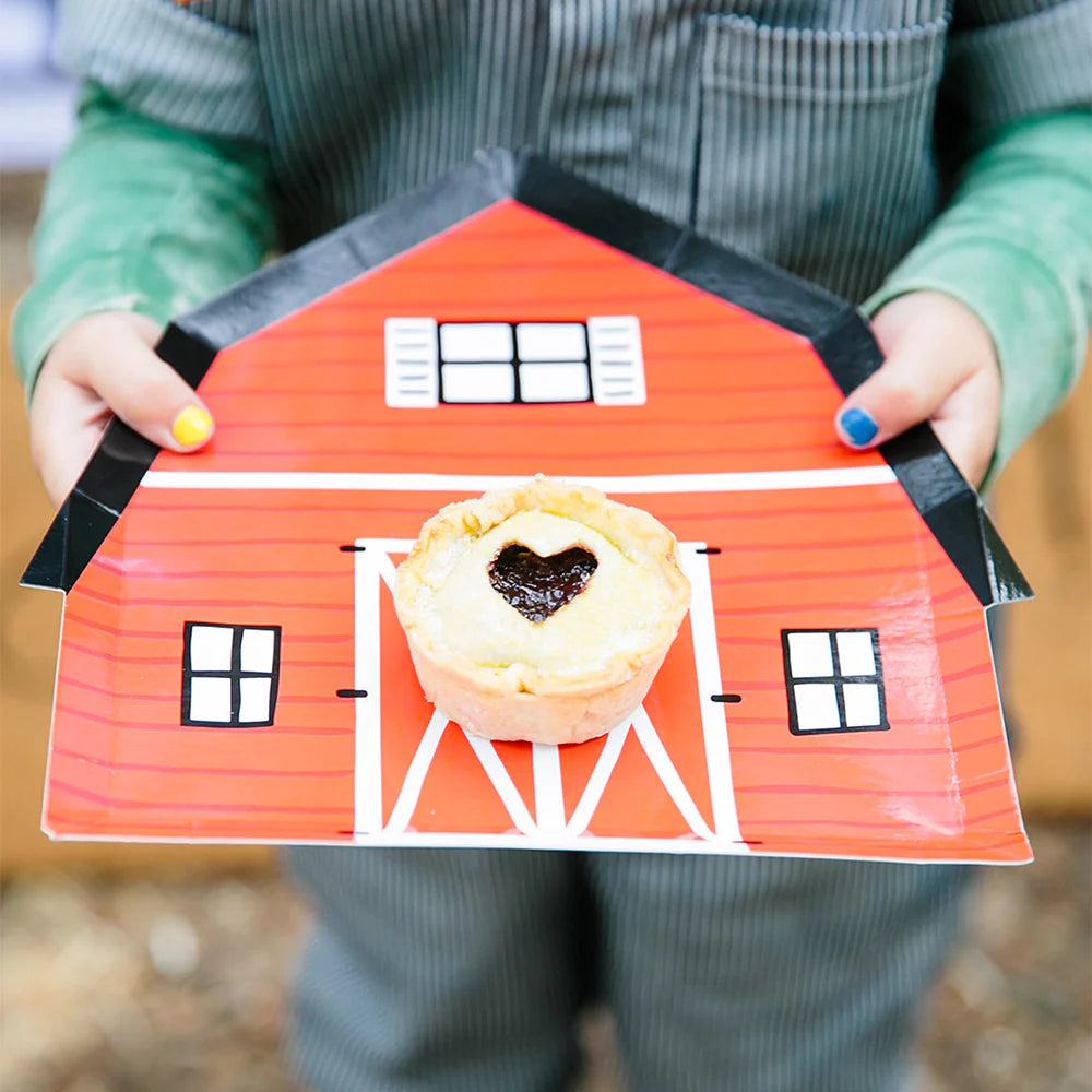 Child holding red barn‑shaped plate with mini pie on top—nail‑polished fingers visible.