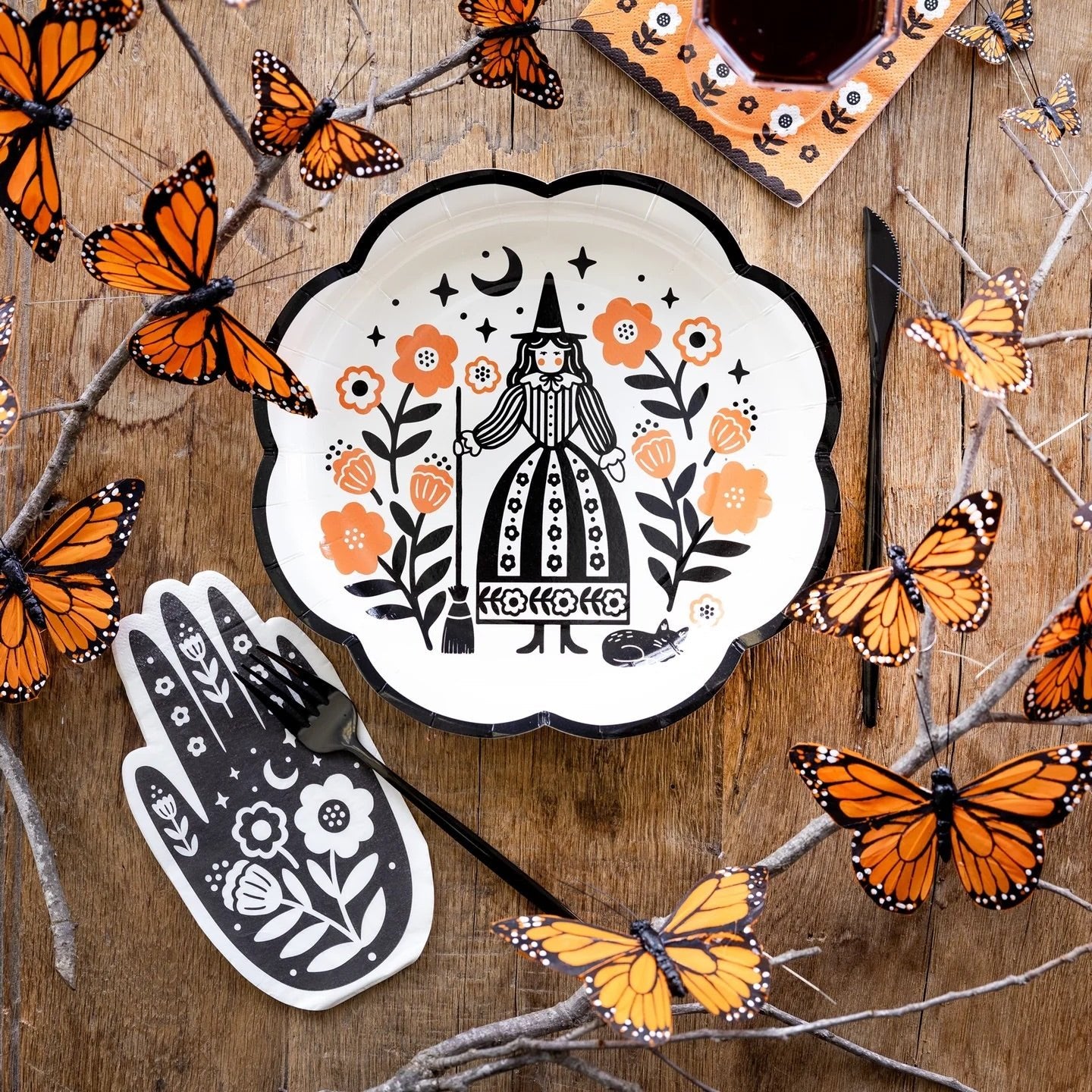Halloween-themed table setup with a witch illustration plate, hand-shaped Halloween paper napkins, and orange floral napkins, accented by orange butterflies for a festive look.