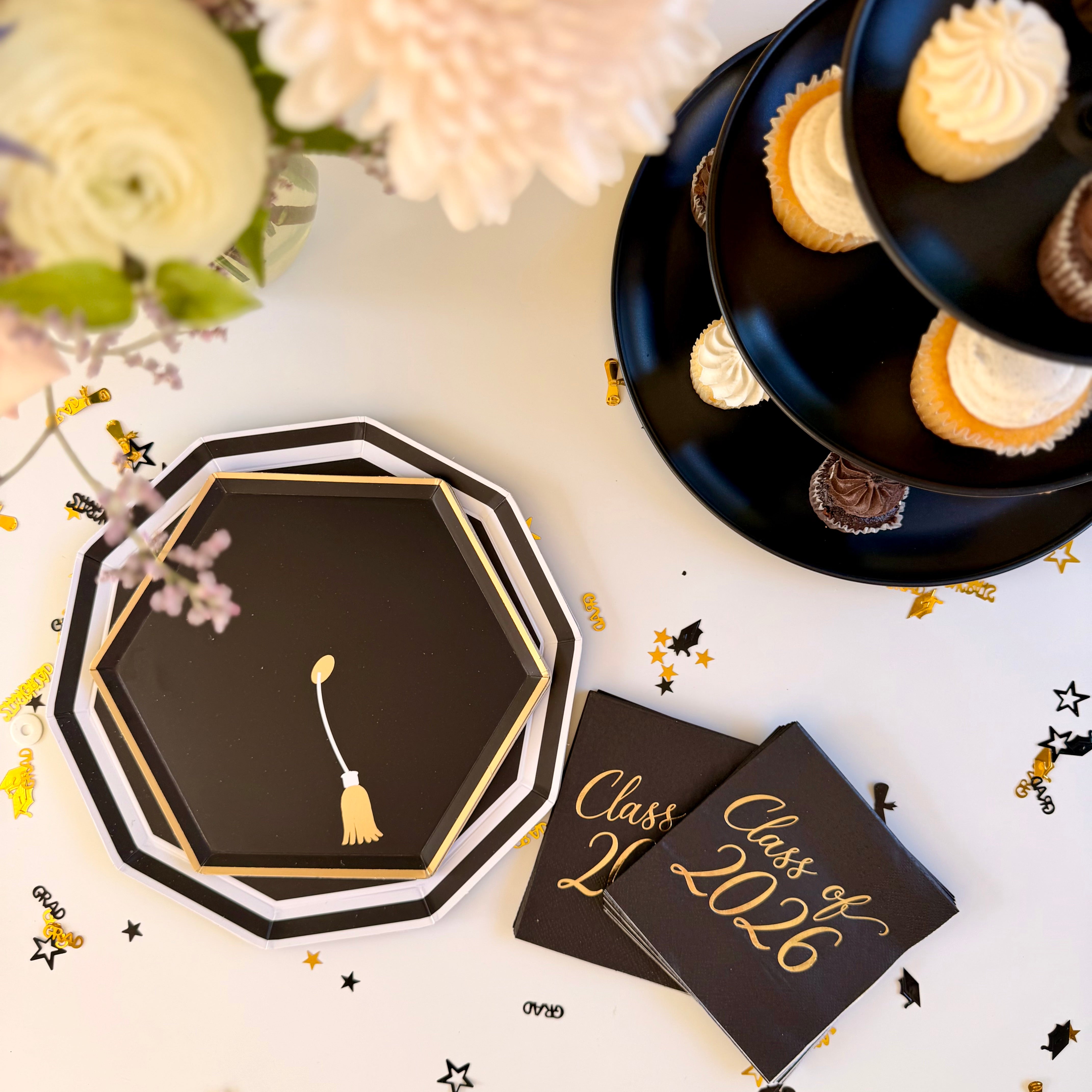 Overhead view of black and gold grad cap paper plates and Class of 2026 cocktail napkins on a white table, styled beside a tiered stand of vanilla and chocolate cupcakes