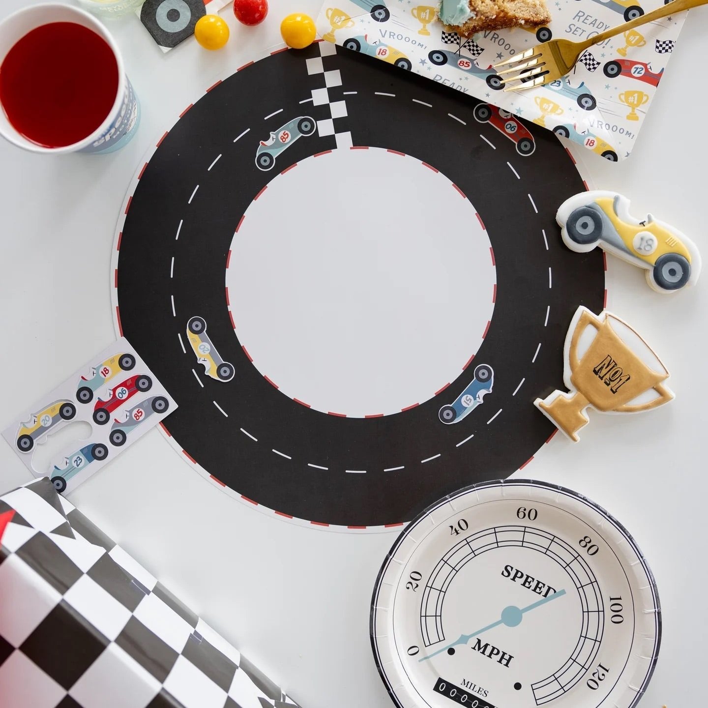 Overhead view of speedometer plates on a race track table setting with colorful car-themed party decor and themed snacks.
