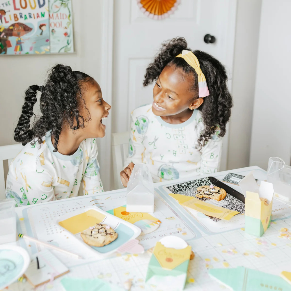 Two kids laughing at a decorated table with crayon box napkins, notebooks, and snacks, set for a back to school celebration.
