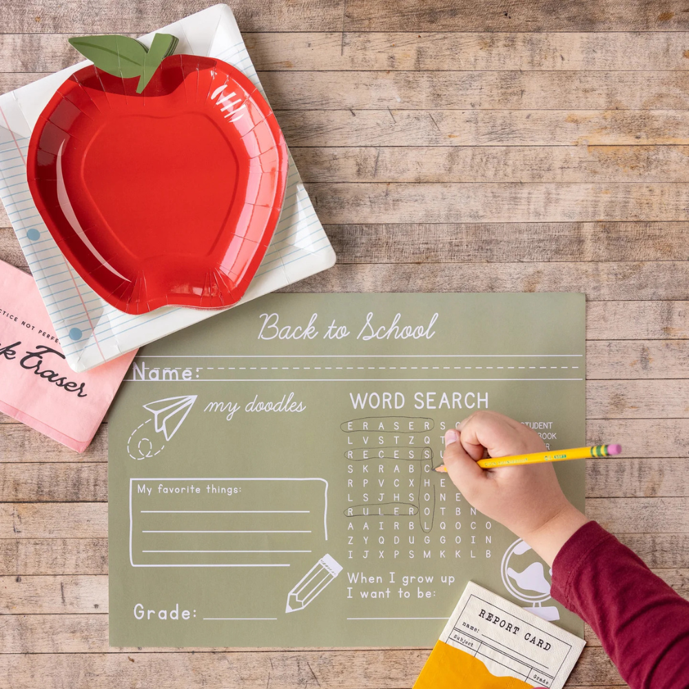 Flat lay of a table setup featuring a green school-themed placemat, red apple plate, and pink eraser-shaped napkin next to a child doing a word search.