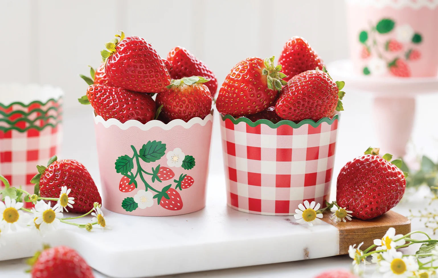 Close-up lifestyle image of two baking cups—one pink with strawberries and one gingham—both filled with juicy strawberries on a white marble countertop.