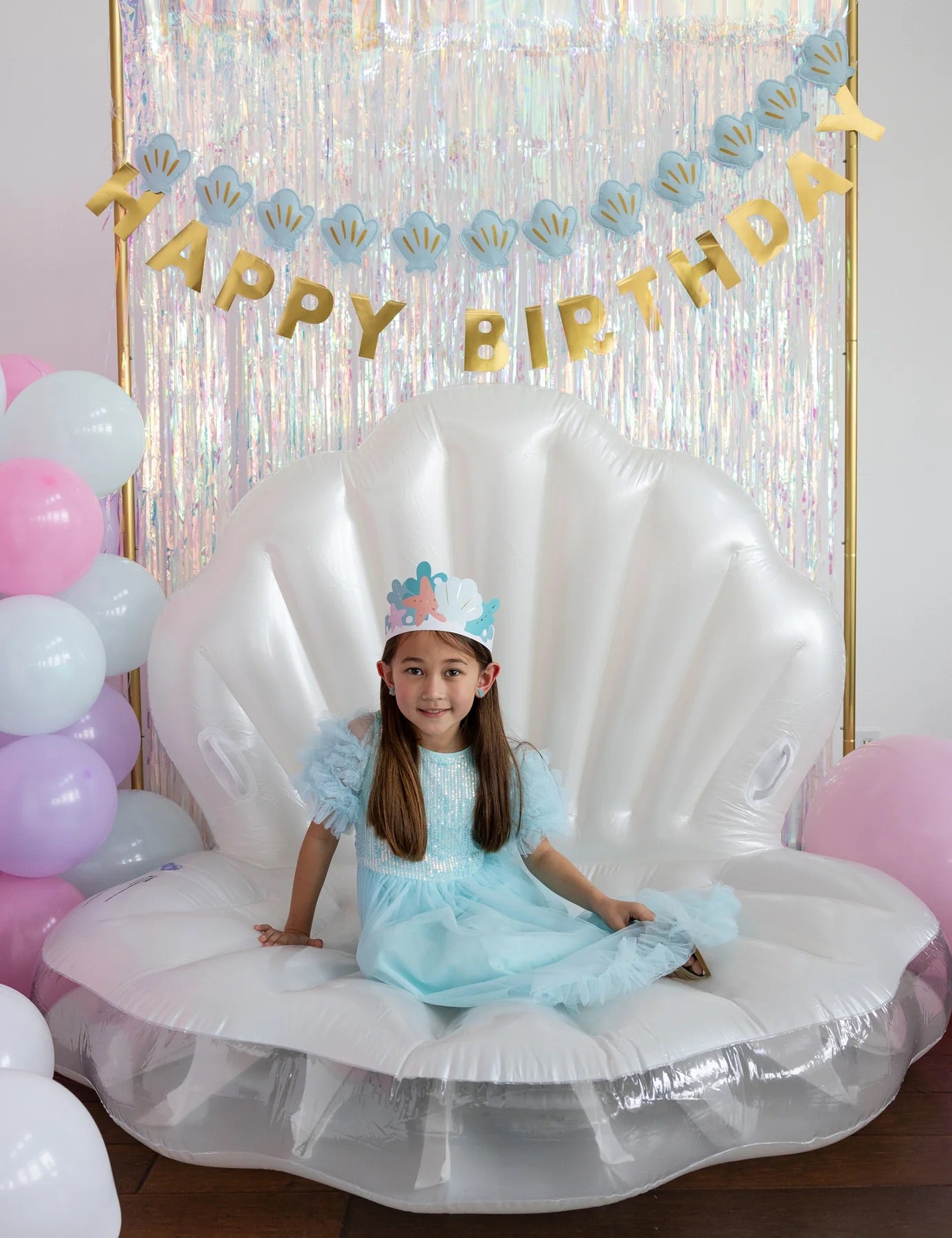 Vertical image of girl wearing mermaid crown seated on large inflatable shell throne, surrounded by iridescent streamers and birthday banner.