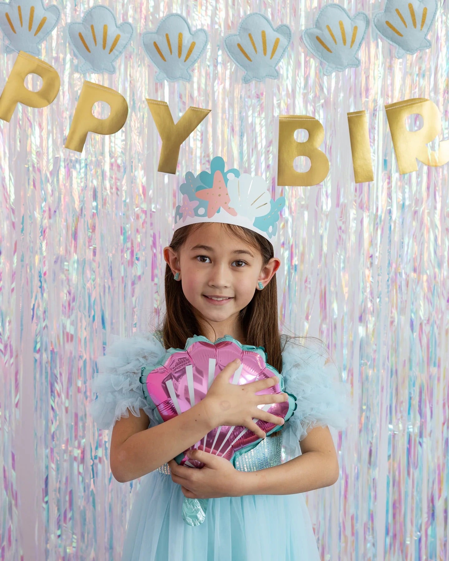Vertical image of smiling girl wearing pastel mermaid crown, holding pink seashell balloon, standing against iridescent background with birthday decorations.
