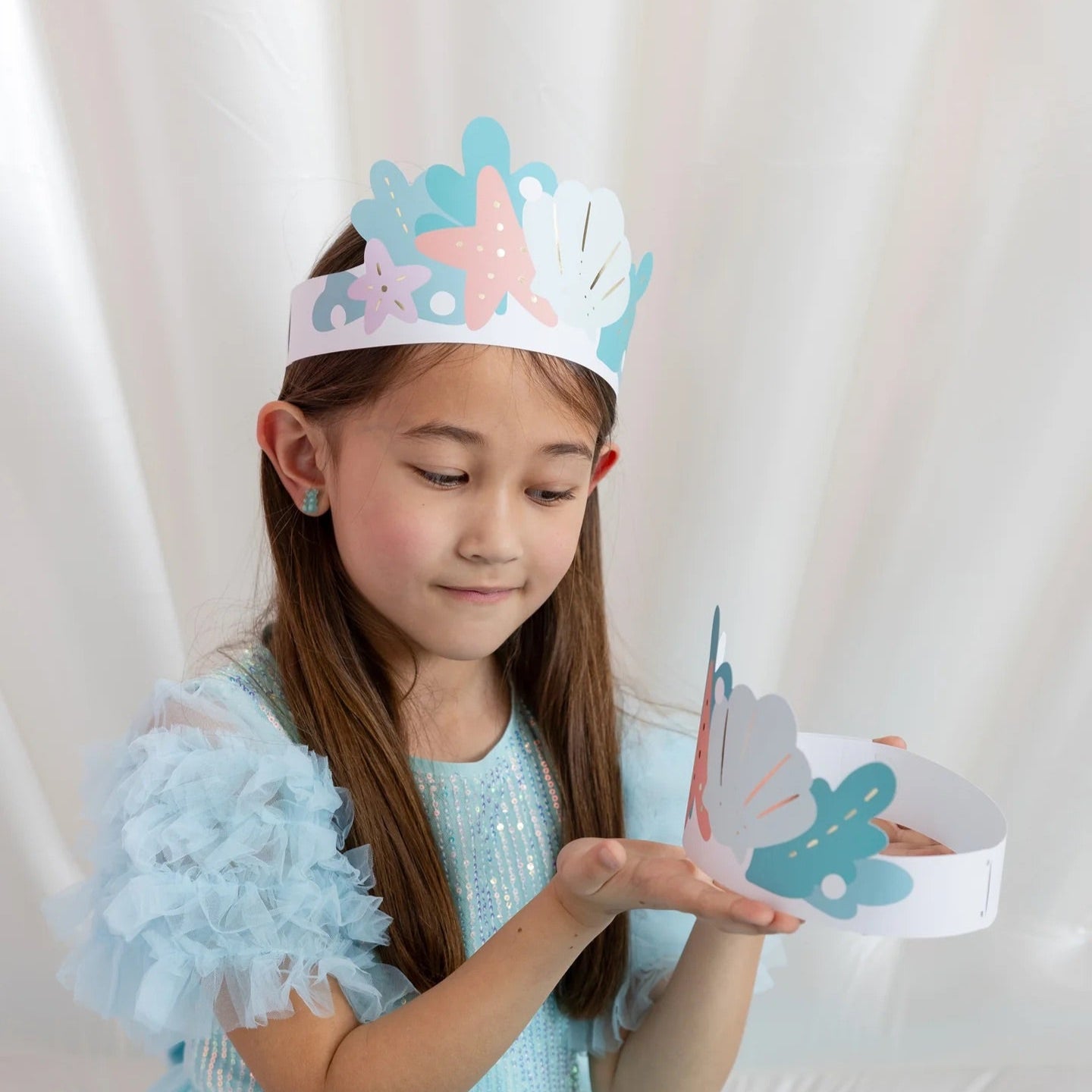 Sweet image of girl wearing a mermaid crown and holding another crown, enjoying her mermaid-themed birthday.