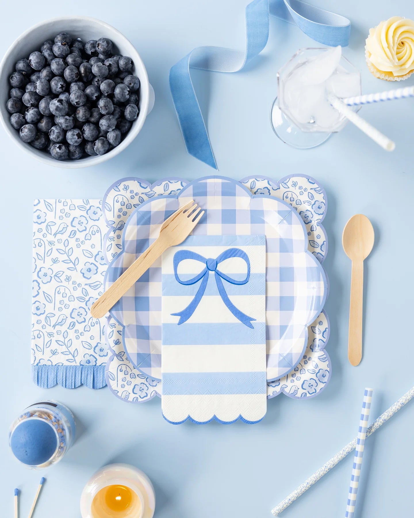 Flat‑lay of a pastel‑blue party setup: layered blue‑gingham and floral scalloped plates with a striped bow napkin and wooden fork, surrounded by a bowl of blueberries, ombré ribbon, iced drink, cupcake, and reusable blue plaid straws on a light‑blue backdrop.
