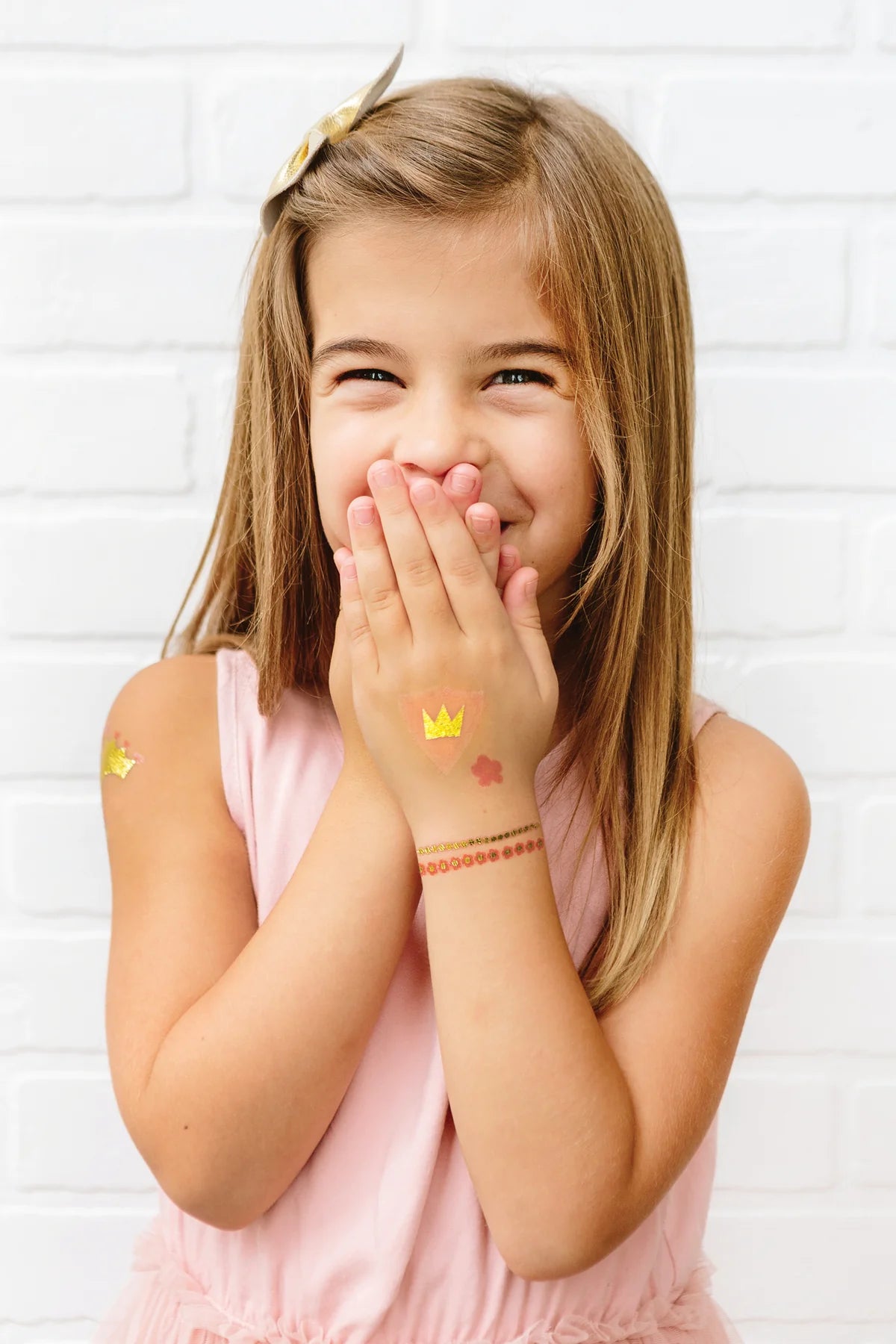 Smiling girl wearing multiple princess tattoos: gold crown on hand, floral bracelet on wrist, small crown on arm.