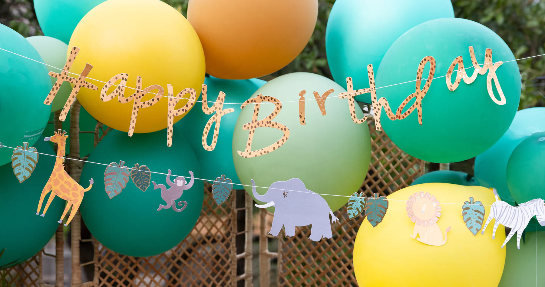 Close-up of a gold foil "Happy Birthday" banner featuring animal print lettering, with a second banner of playful safari animals like a giraffe, monkey, elephant, lion, and zebra, against a backdrop of balloons.