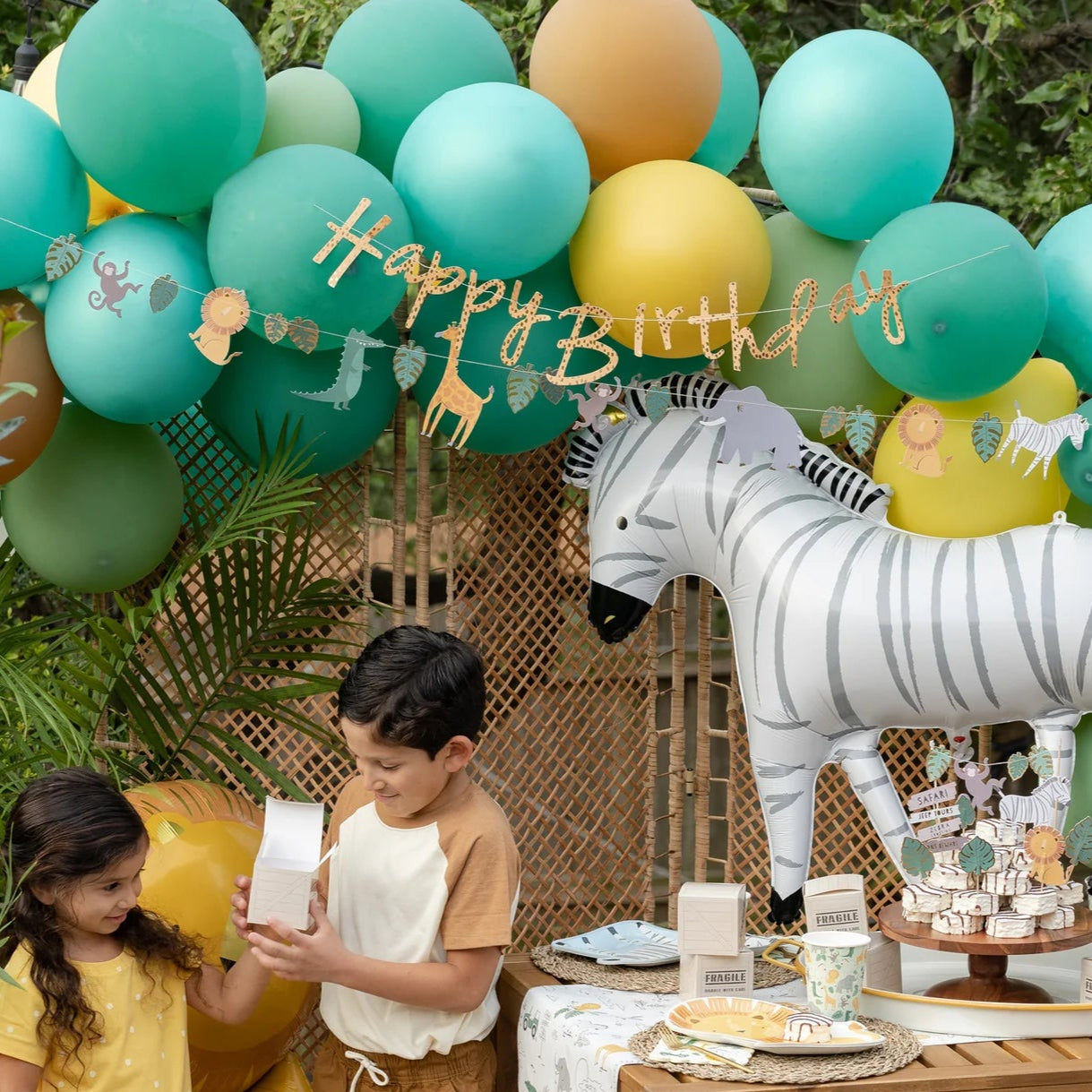  Two smiling children standing by a safari-themed birthday party setup, featuring a large zebra balloon, green and yellow balloon arch, and a festive banner with "Happy Birthday" and safari animals.