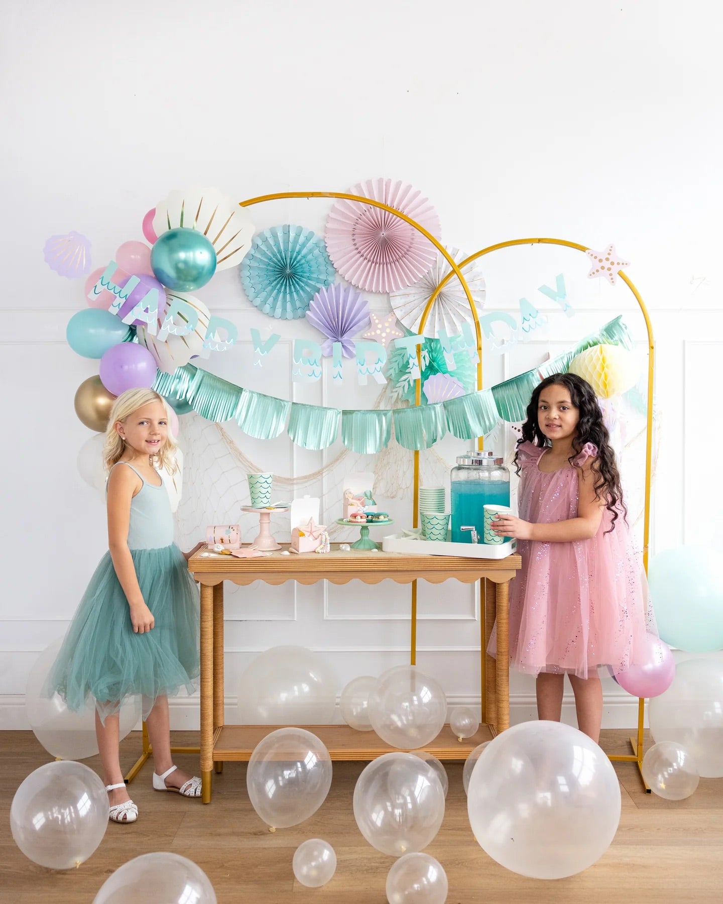 Two smiling young girls dressed for a mermaid party standing near a whimsical dessert table with sea-inspired decorations and pastel-colored balloons.