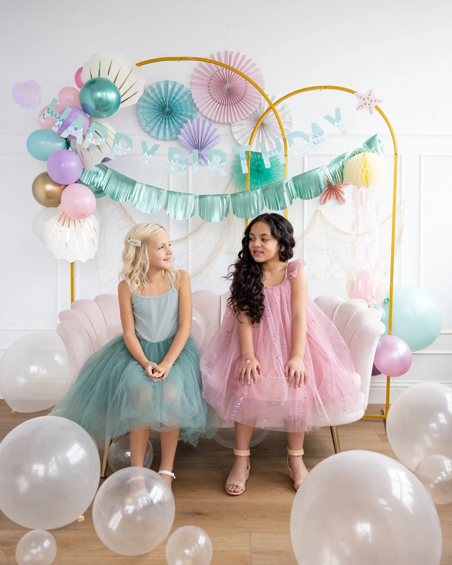 Two little girls in party dresses sitting together beneath vibrant mermaid-themed paper fans and pastel balloon decorations.