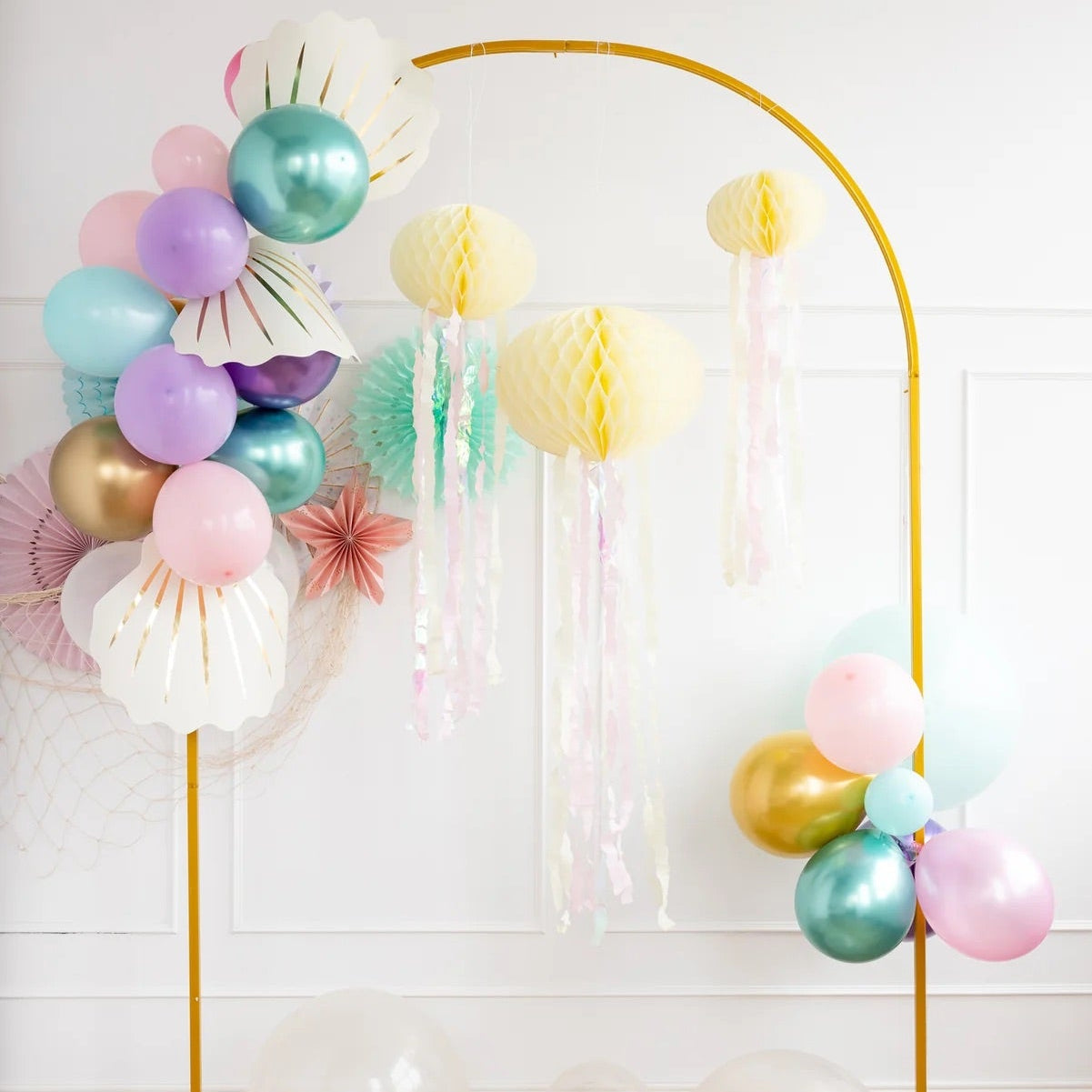 Three pastel-colored honeycomb jellyfish decorations hanging beautifully with shimmering streamers, surrounded by balloons—perfect for an under-the-sea mermaid party.