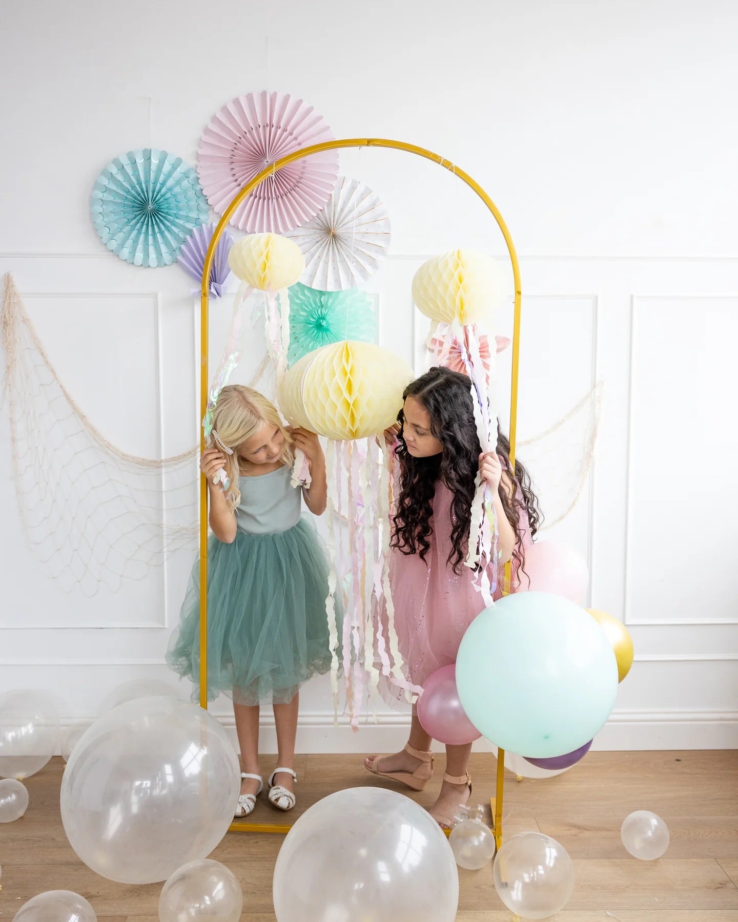 Two girls in party dresses playfully exploring pastel honeycomb jellyfish decor with iridescent streamers, ideal for an enchanting mermaid party.