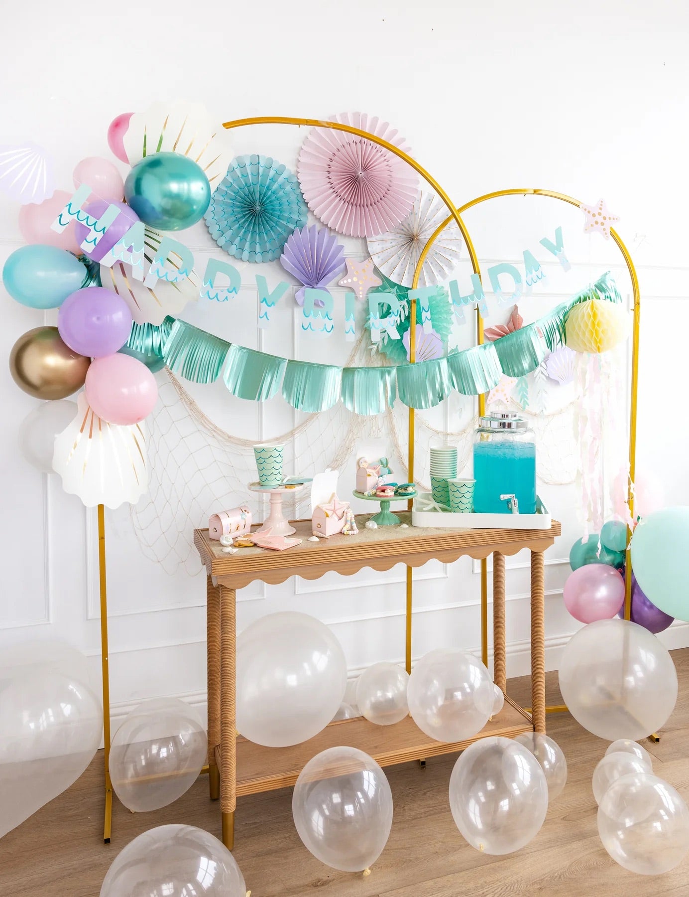 Festive mermaid birthday table setup featuring aqua foil banners, ocean-inspired decorations, pastel balloons, and clear balloons scattered around a wooden serving table.