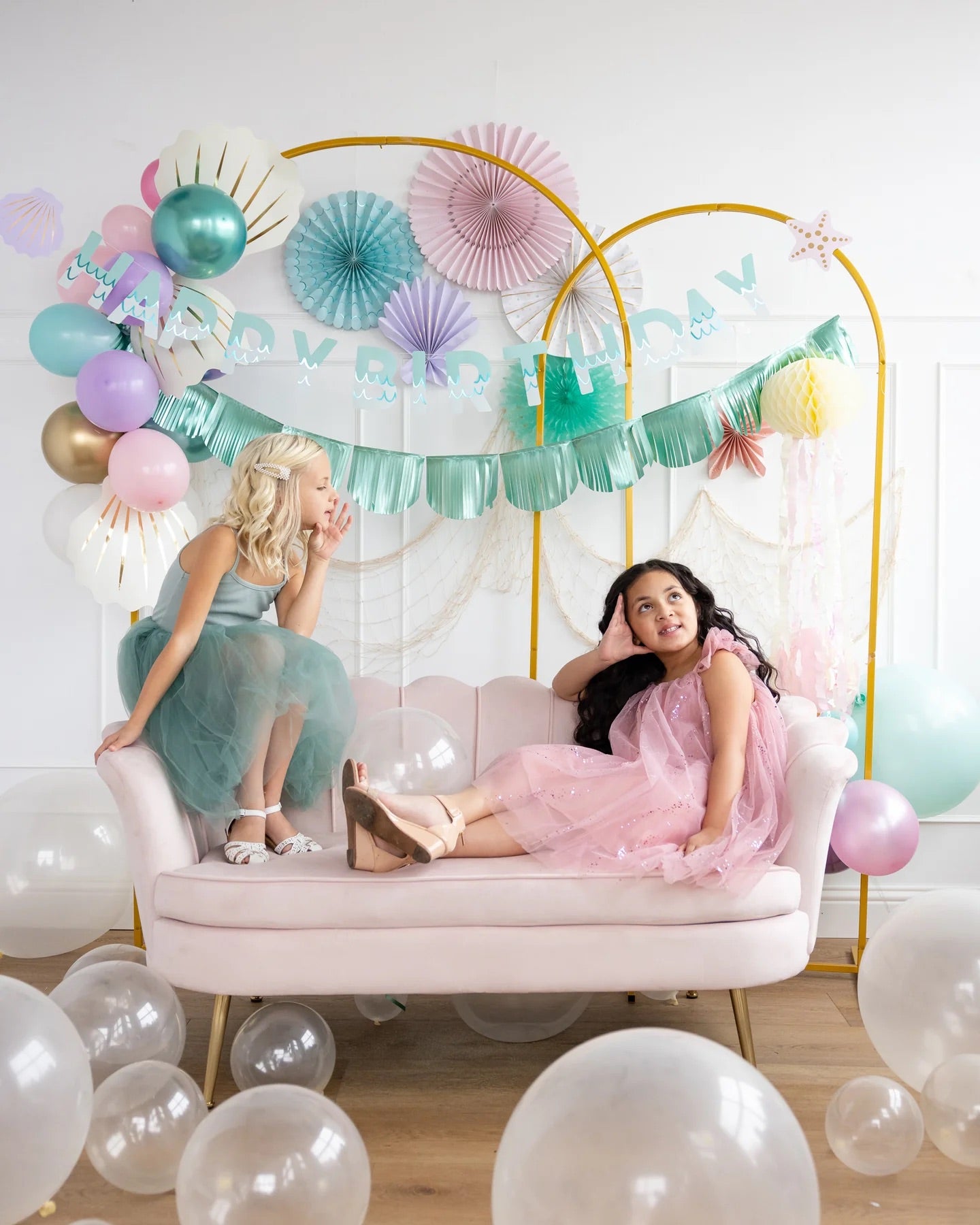 Two young girls lounging and laughing on a pink velvet sofa beneath mermaid-themed birthday decor featuring aqua foil banners, pastel balloons, and ocean-style paper fans.