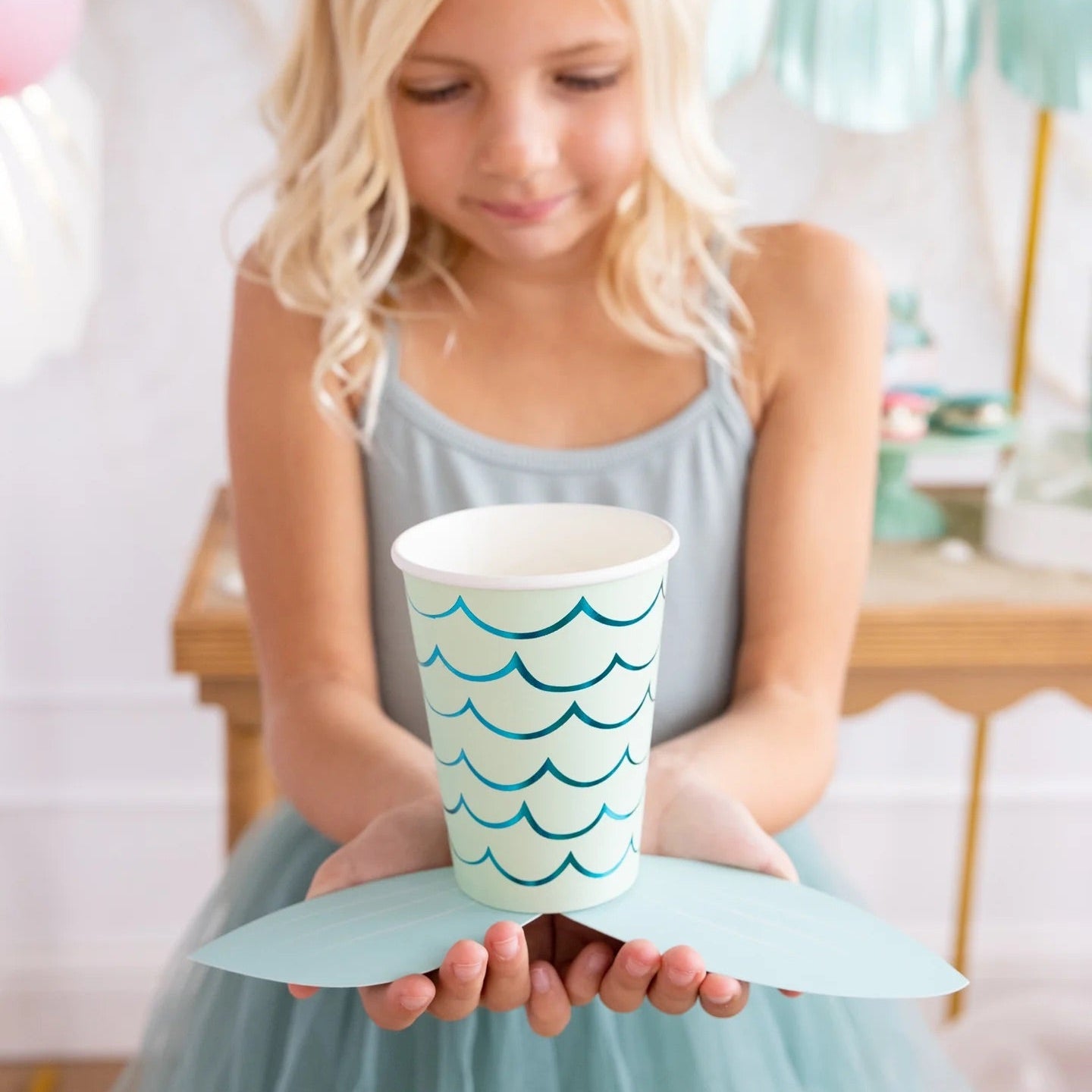 A smiling young girl holding a paper cup decorated with blue foil mermaid scales and a cute aqua adhesive tail.