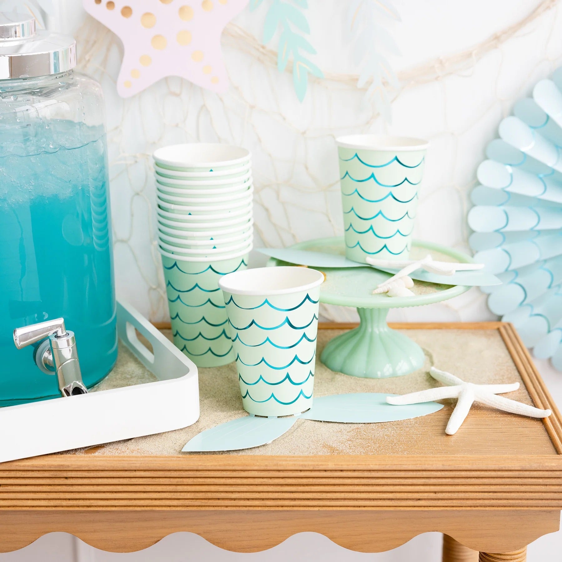 Party display table featuring stacked mermaid-themed paper cups, starfish decor, and a dispenser with blue drink, perfect for a mermaid birthday celebration.