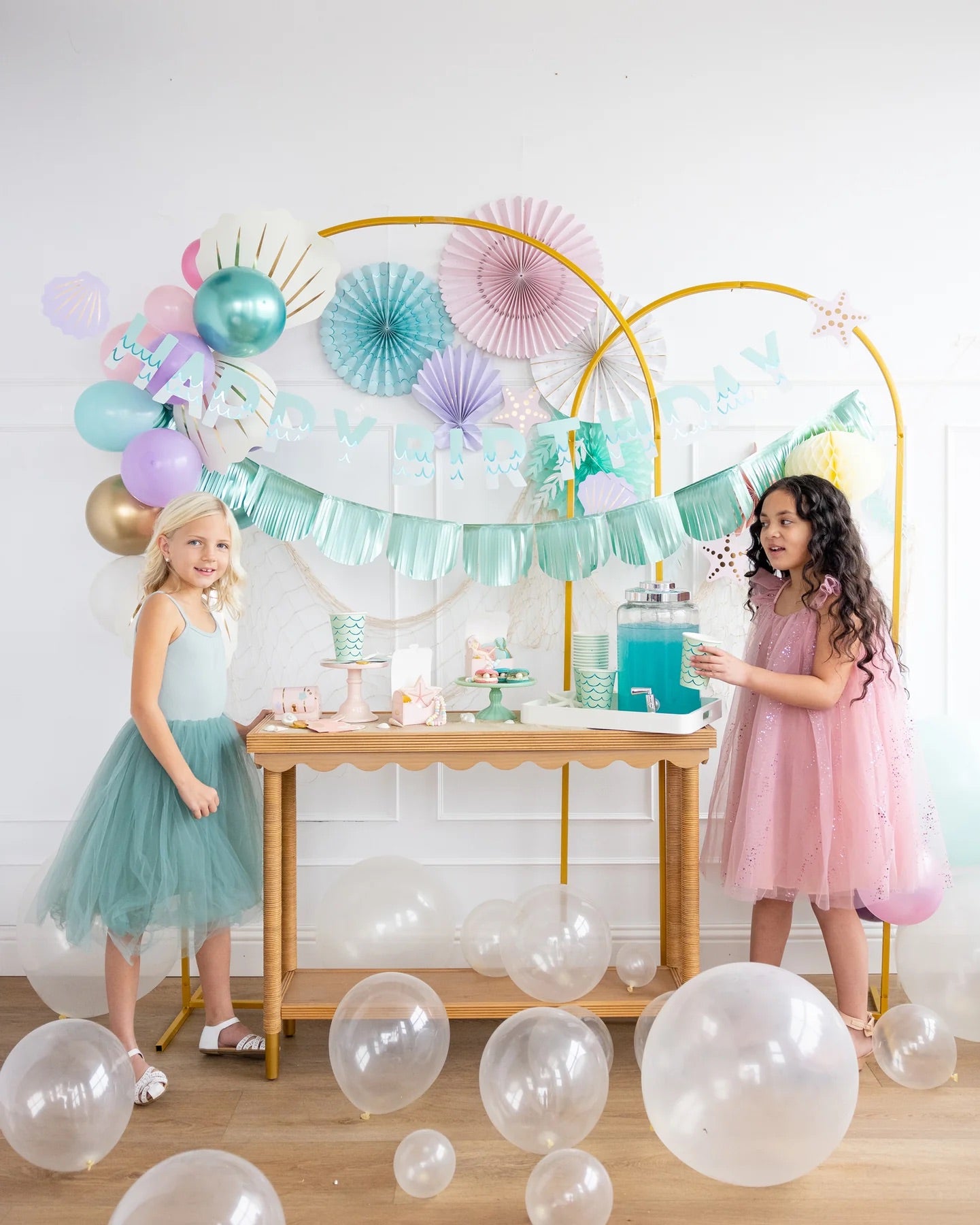 Two young girls, smiling, at a mermaid-themed party table with decorations, balloons, and mermaid cups.