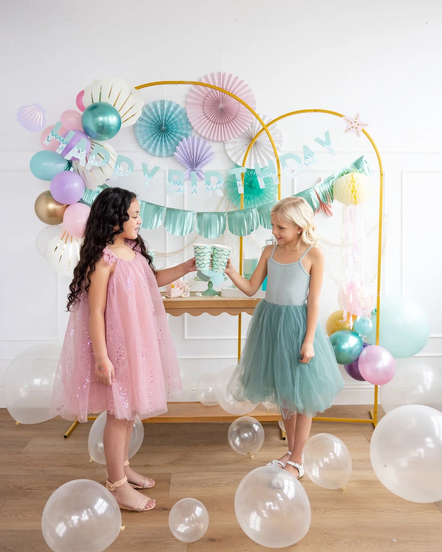 Two girls dressed in pastel party dresses clinking mermaid-themed cups, standing in front of a mermaid-themed birthday banner with balloons and decorations.