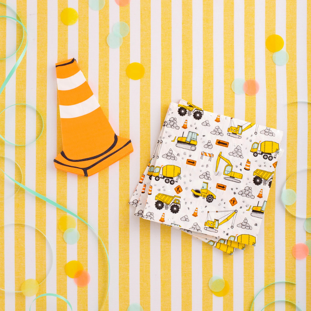 Construction-themed party table with yellow striped tablecloth, traffic cone-shaped napkin, and folded large napkins covered in illustrated construction vehicles and signs.
