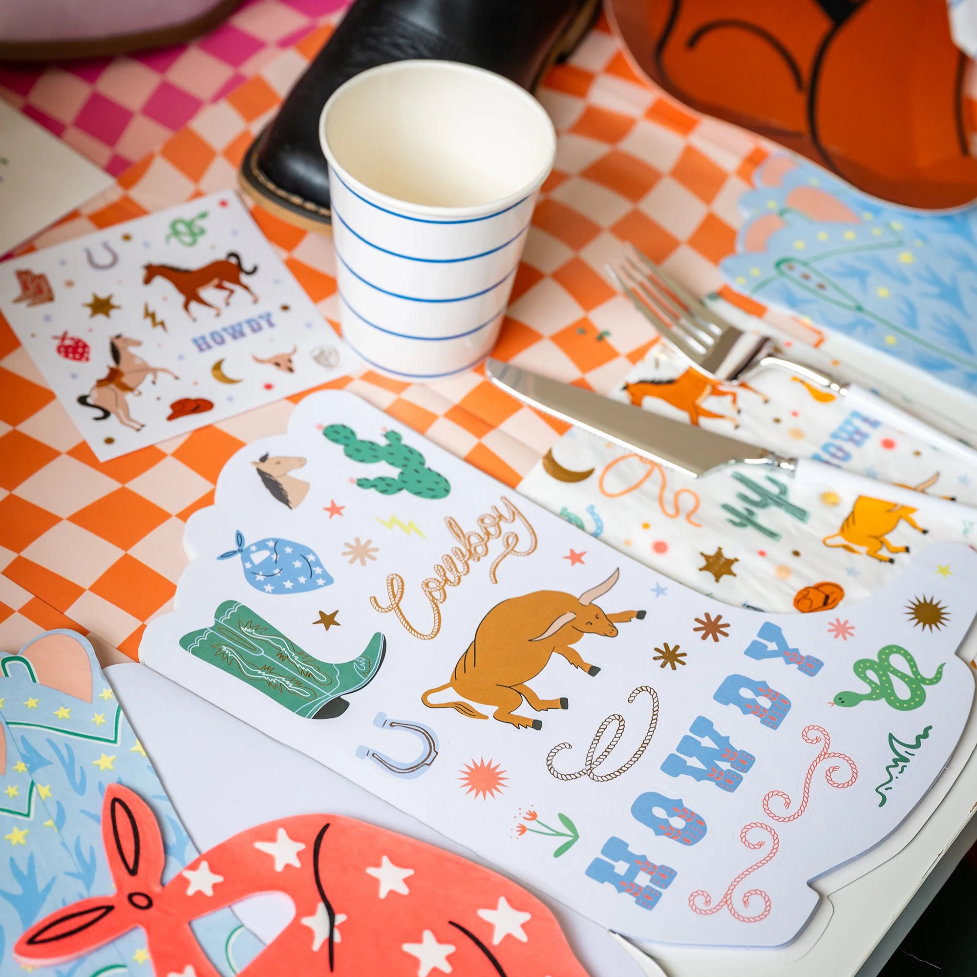 Sticker sheet on orange checker table: Close‑up of stickered sketchbook page next to striped cup and place setting on orange gingham tablecloth.