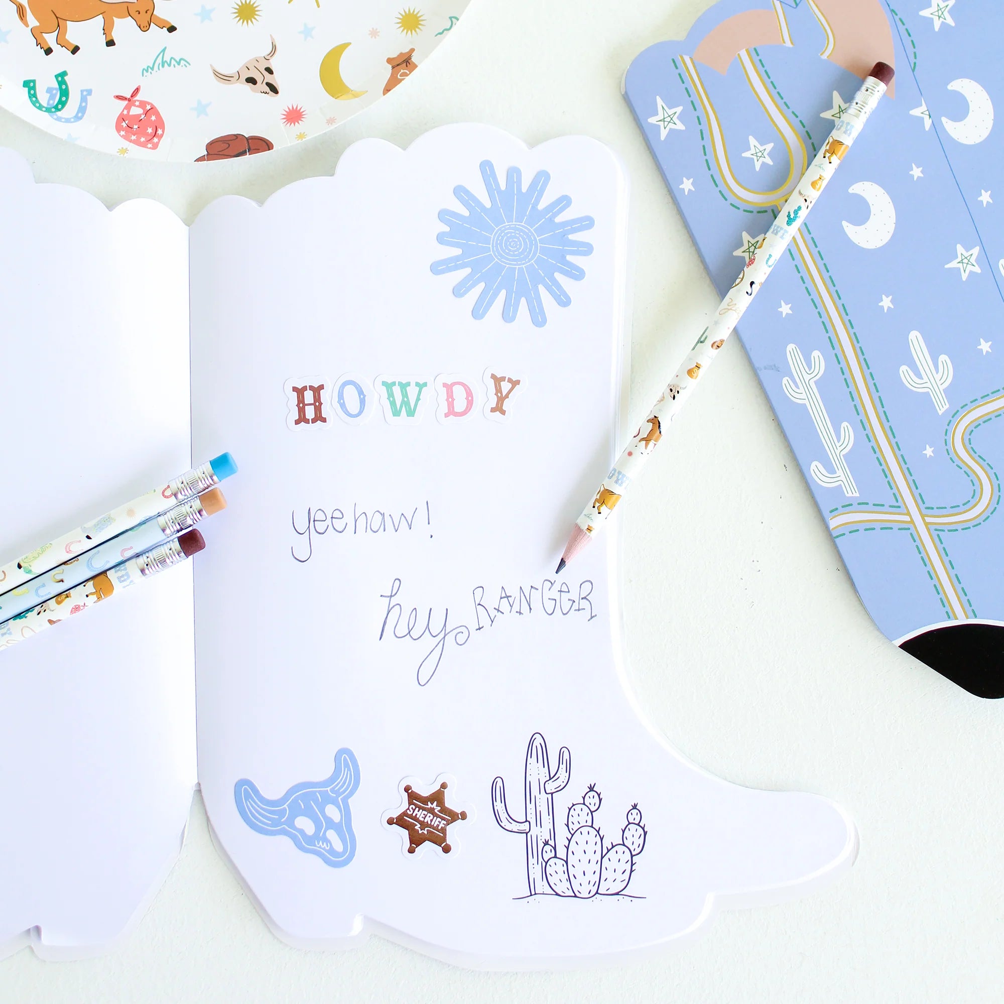 Open book spread: Boot‑shaped sketchbook opened flat with blank white pages, western stickers, and handwritten “Howdy” doodles alongside patterned pencils.