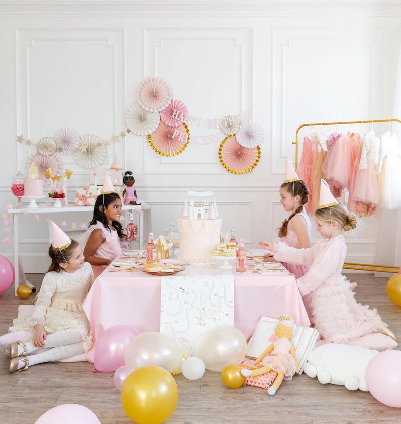 Wide shot of pink birthday table, dress-up rack of tulle gowns, and gold-rim fans hanging as elegant princess party decorations.