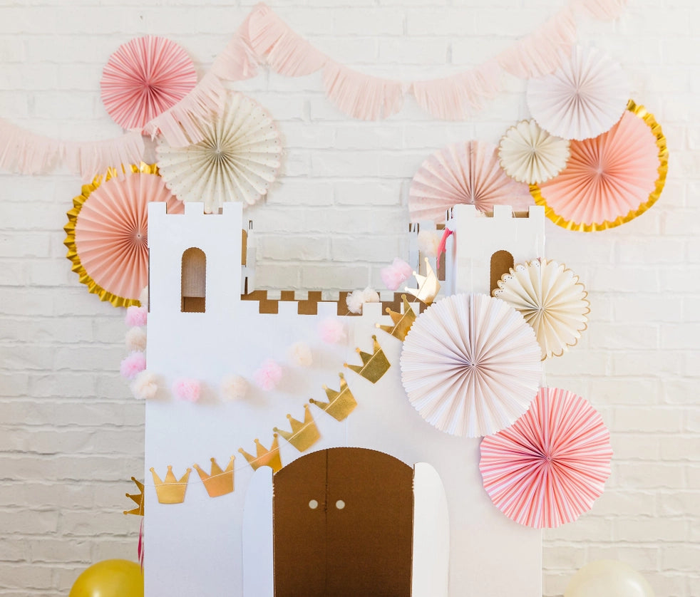 White cardboard castle framed by gold crown and tulle pom pink banner with blush paper fans—ready for a princess birthday party photo wall.