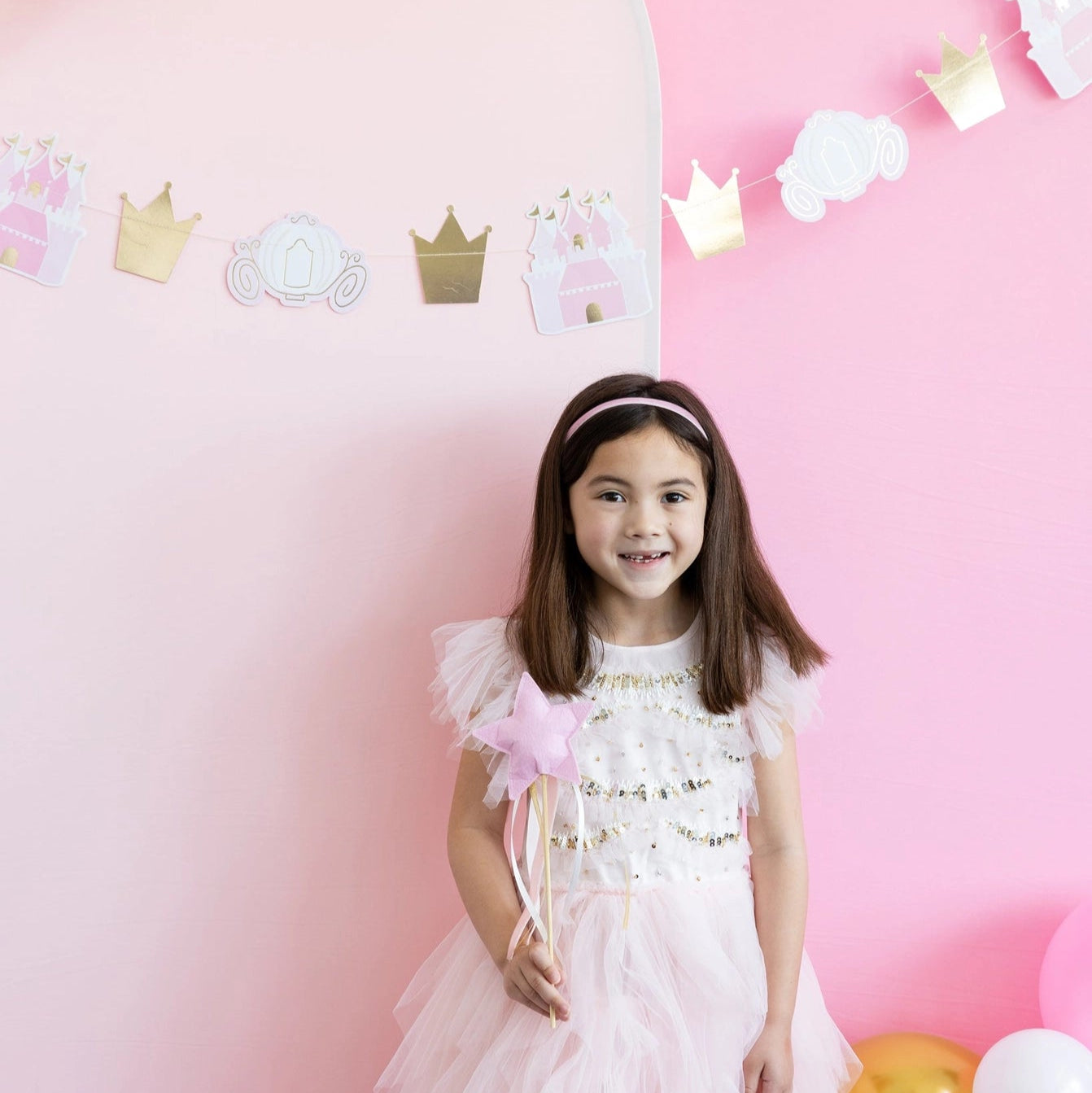 Smiling child posing against pink backdrop while gripping felt star princess wand with trailing ribbons.