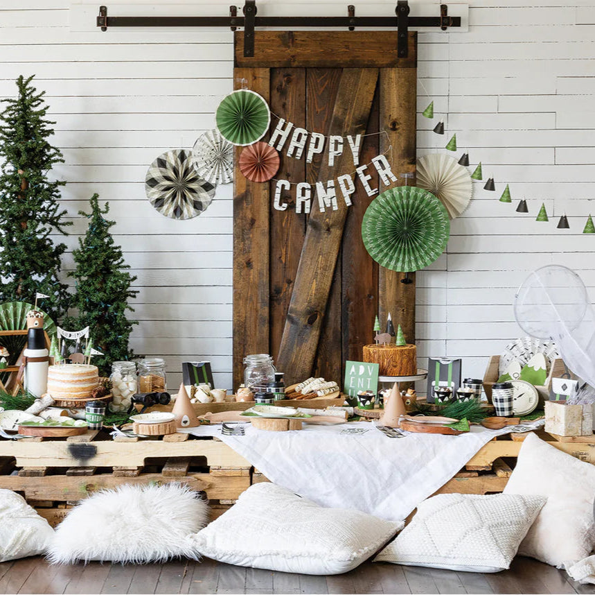 A styled kids' camp birthday party scene with teepees, rustic wood decor, paper fans, and a cozy picnic setup under “Happy Camper” signage.