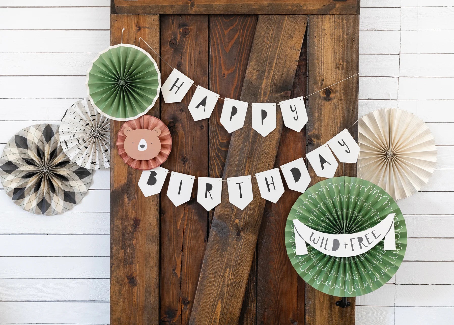 “Happy Birthday” banner hanging on wooden barn doors surrounded by green, plaid, and kraft paper fans for a cozy camping birthday party vibe.