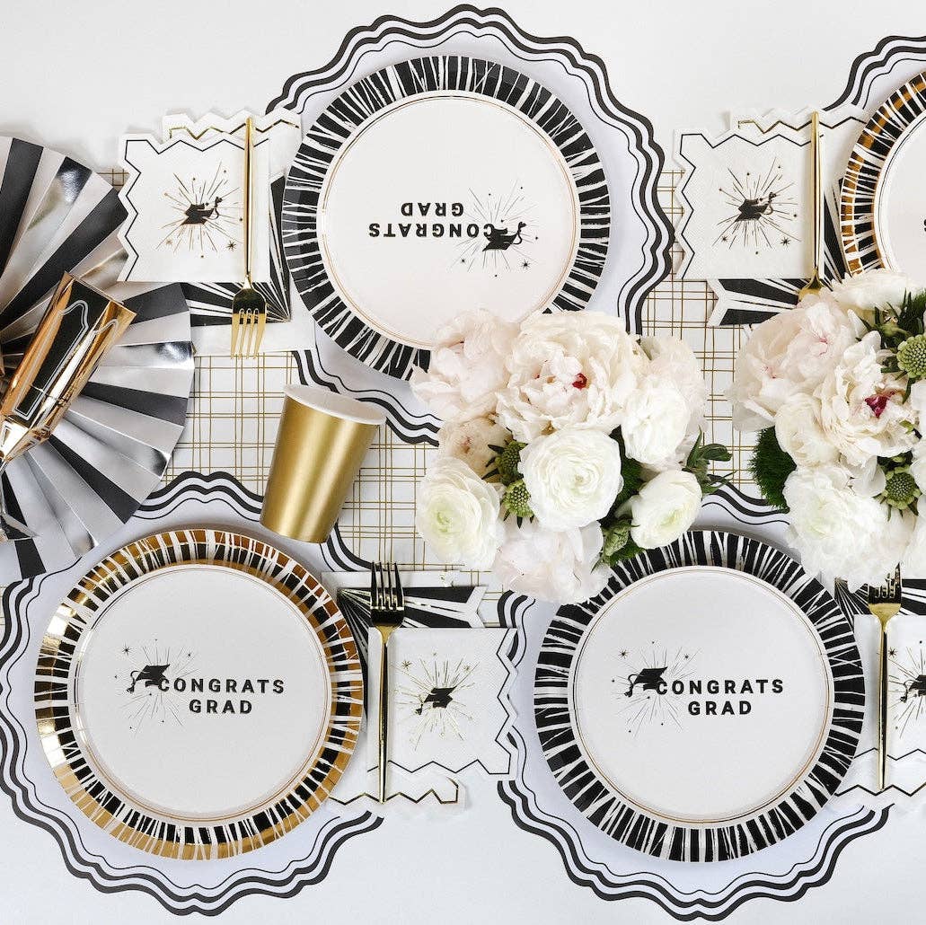 An overhead view of a graduation tablescape with Congrats Grad plates, gold cups, and white peonies.
