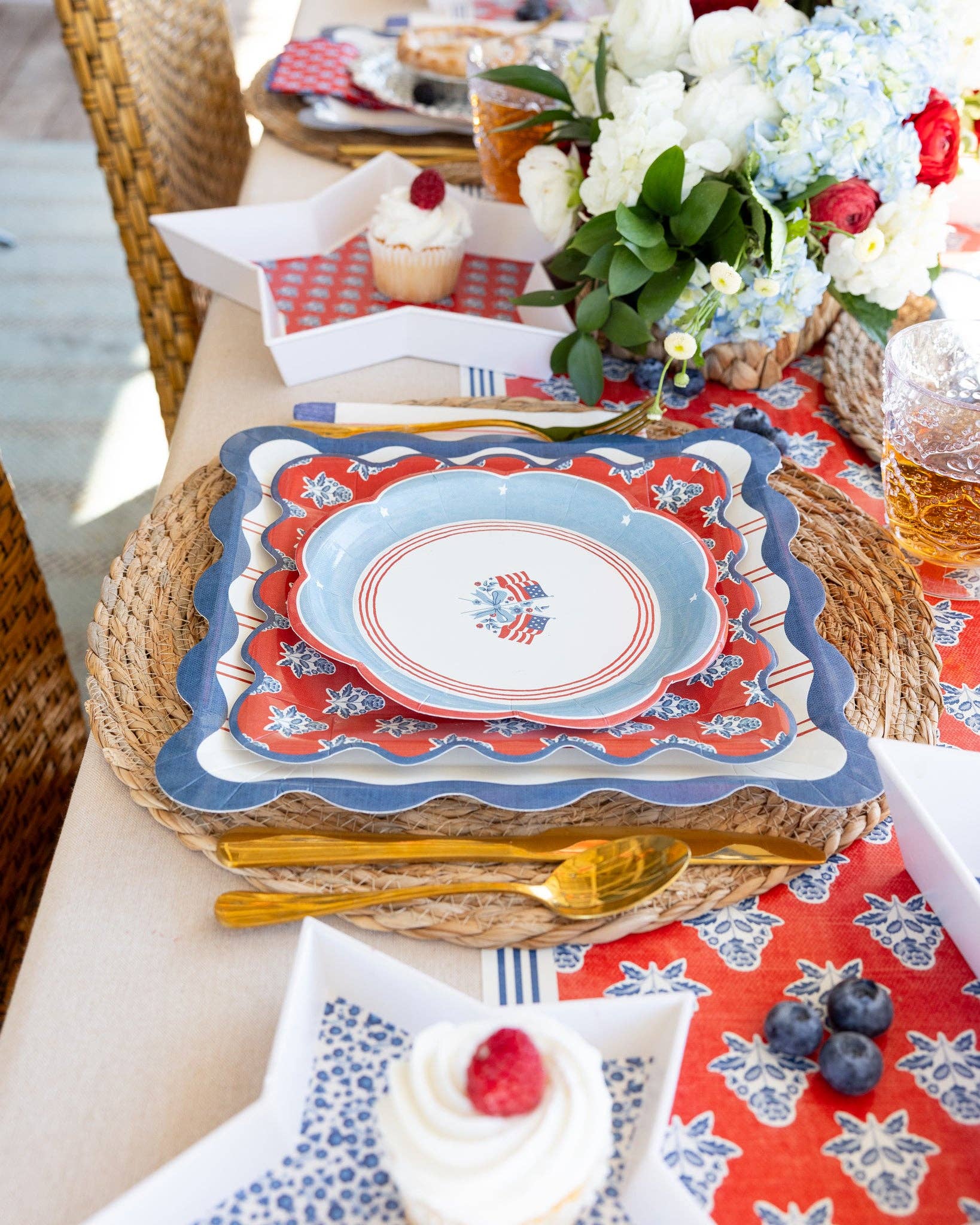 American flag scalloped paper plate layered on red block-print floral plate and red striped dinner plate with wicker placemat, gold cutlery, and Hamptons Americana table setting