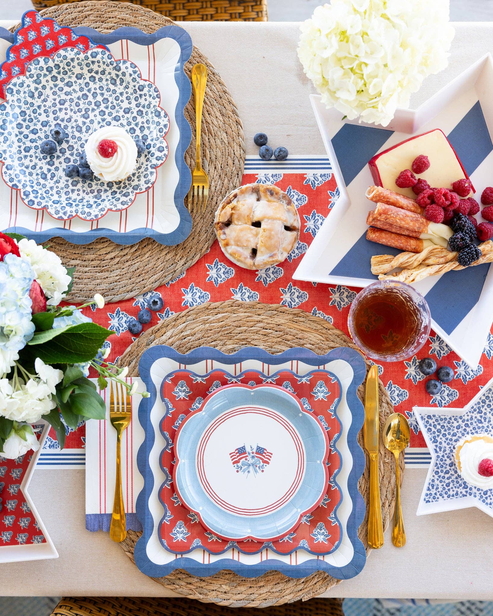 Hamptons Americana full tablescape overhead with American flag scalloped plates, blue ditsy floral plates, red striped dinner plates, red block-print napkins, star trays, hydrangeas, and gold cutlery