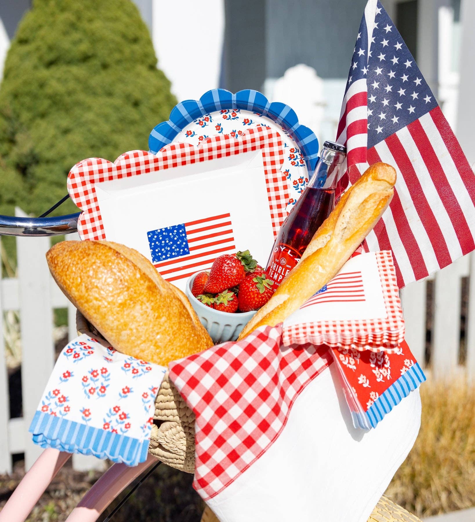 Bicycle basket styled with American flag cocktail napkins, Americana paper plates, strawberries, baguettes, and an American flag in a summer picnic scene
