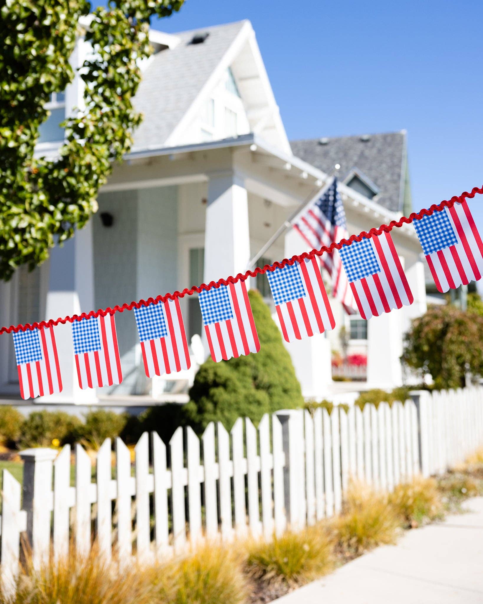 Close-up of the Americana flag banner hanging across a white picket fence in front of a white house with an American flag on the porch