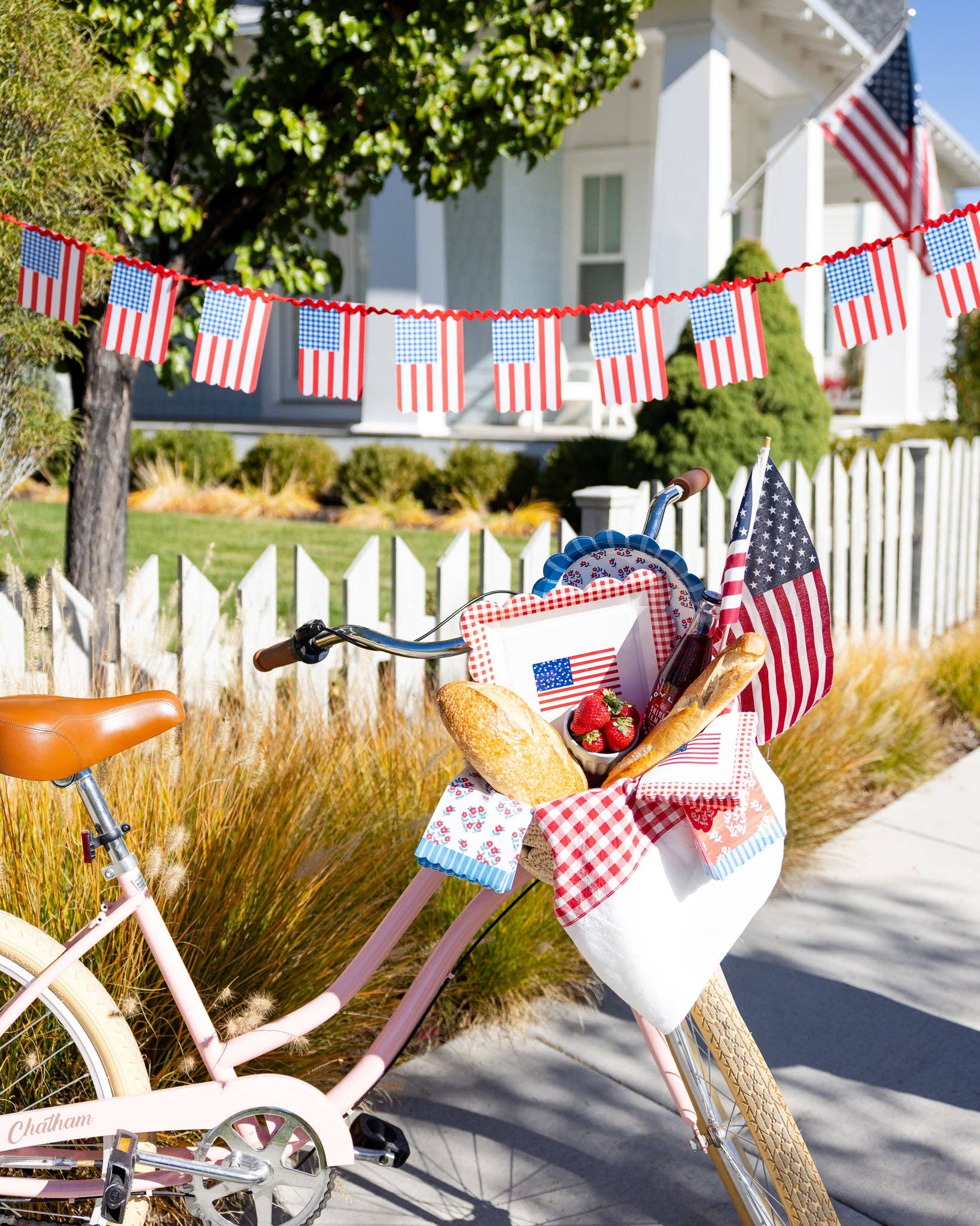 Pink bicycle with basket filled with gingham flag plates, floral scalloped plates, floral napkins, and American flags in front of white picket fence