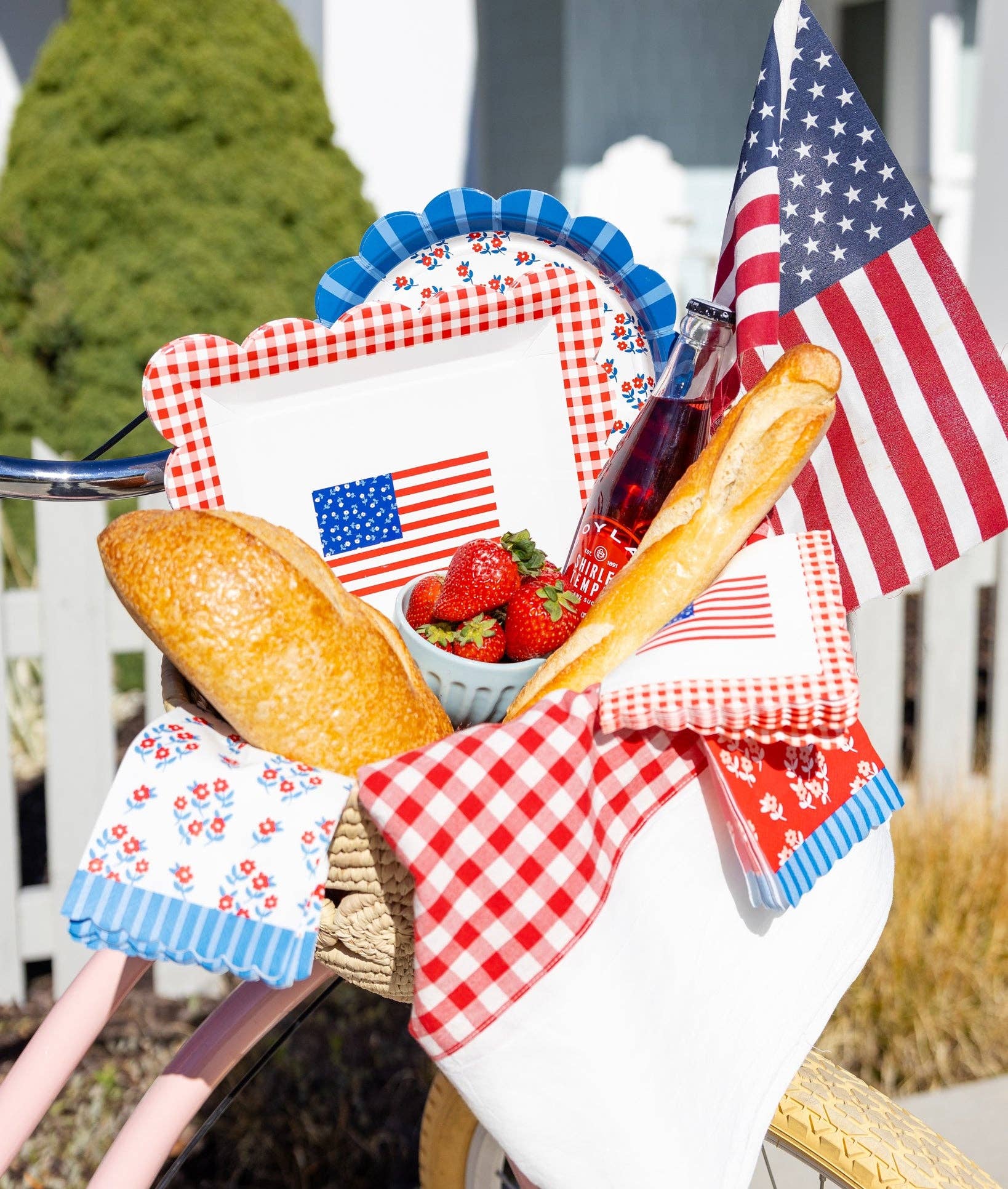 Close-up of pink bicycle basket with gingham flag plates, floral scalloped plate, strawberries, baguettes, and American flags