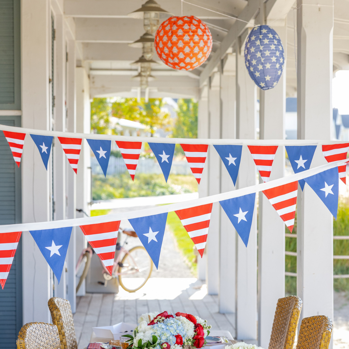 Red white and blue star pennant bunting strung across white porch with Americana paper lanterns hanging above outdoor tablescape